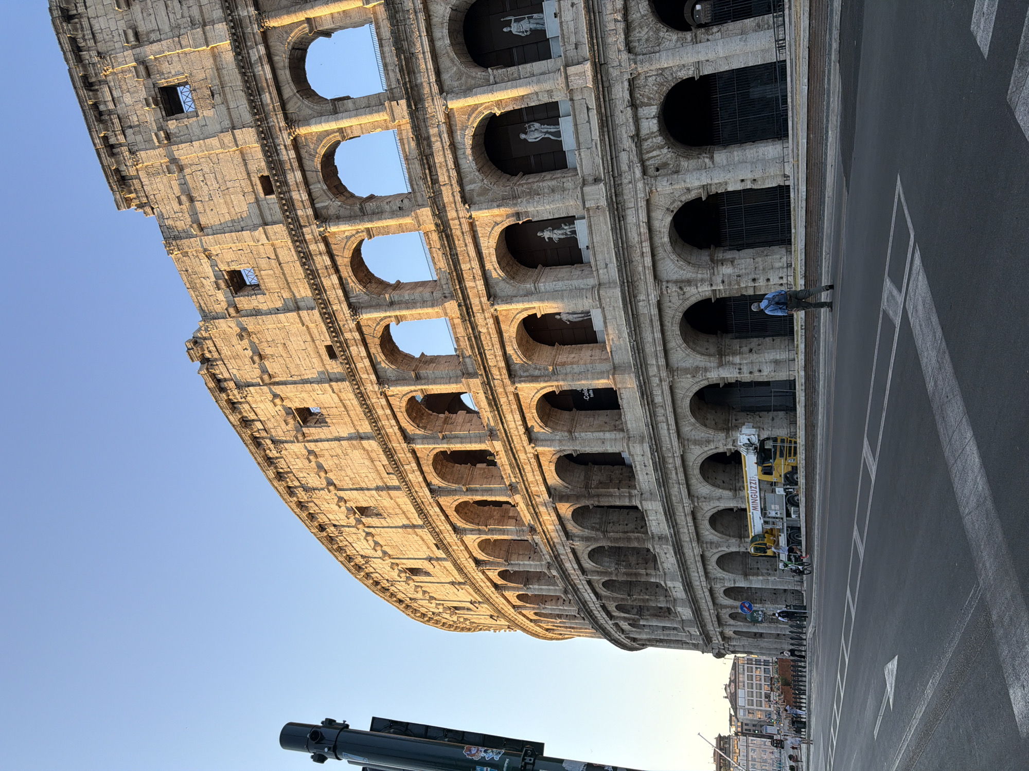 The image shows the Colosseum in Rome, Italy, under a clear blue sky. The sun illuminates the ancient amphitheater, casting long shadows and highlighting the textured stone. In the foreground, a street with white lane markings and a pedestrian crossing is visible. A few people are present: one person walks away from the camera on the street, wearing a blue shirt and backpack; another person is on a bicycle near a yellow crane truck with the name "MINGUZZI" on it. Several other individuals are seen walking or standing further away along the sidewalk in front of the Colosseum. The lower arches of the Colosseum are closed off with dark metal gates, while higher up, some of the openings feature statues. The overall scene suggests a quiet morning or late afternoon in a tourist area.