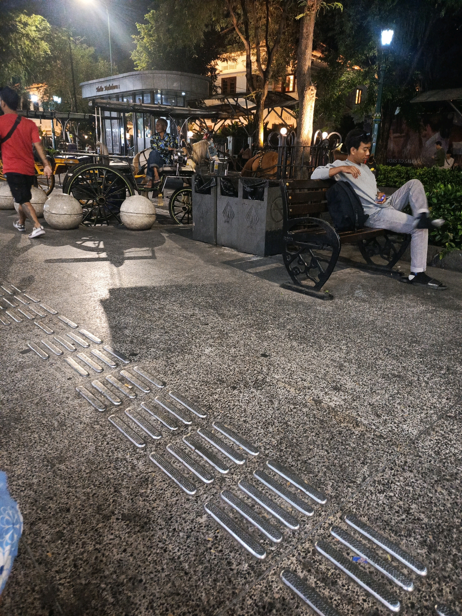 The image depicts a street scene in Yogyakarta, Indonesia, likely near a tourist area given the presence of horse-drawn carts, known locally as "andong." In the foreground, a textured pavement with tactile paving stones for the visually impaired is visible. A young man, casually dressed in a gray long-sleeved shirt and light-colored pants, is seated on a dark wooden bench on the right side of the frame, leaning back with his legs crossed and holding a small object. He is looking towards the left. Behind him, a dark backpack rests on the bench.
To the left, a person in a red shirt and black shorts is walking away from the camera, casting a shadow on the ground. In the mid-ground, several horse-drawn carts are parked. One cart has a man dressed in a colorful shirt seated in the driver's position, holding reins. Another person is visible within this cart. Large, spherical concrete bollards are placed in front of the carts. The background reveals trees, streetlights, and buildings, suggesting an urban setting at dusk or night, illuminated by artificial light. The overall atmosphere is that of a lively public space.
