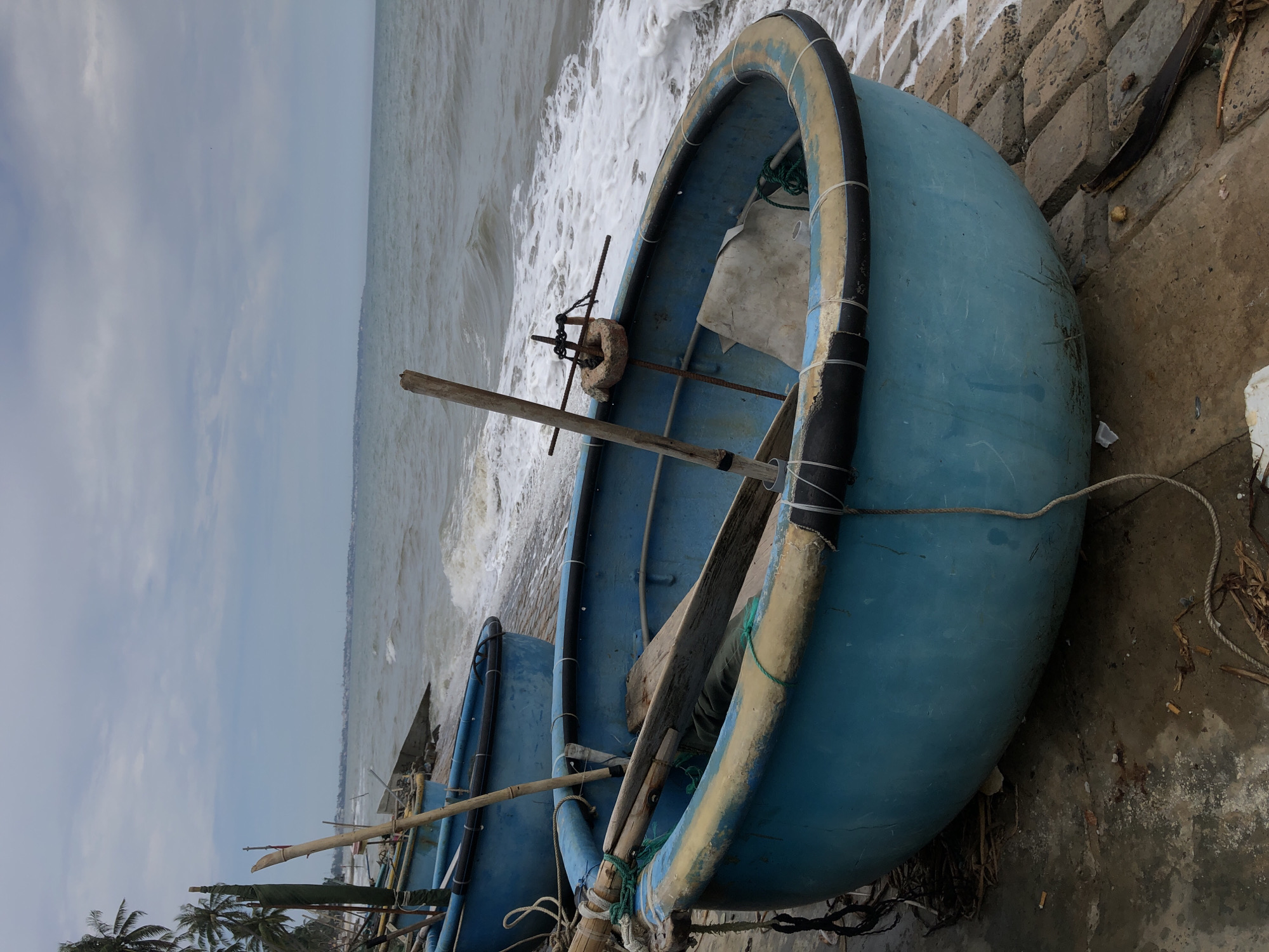 The image shows a close-up of traditional Vietnamese basket boats, known as coracles, pulled up on the shore in Mũi Né, Vietnam. The boats are round, made of woven material, and coated with a blue, waterproof substance. The boat in the foreground is partially filled with wooden oars and other debris. The waves of the ocean are visible in the background, crashing against the shore with white foam. The sky is overcast with clouds. In the far background, palm trees and a distant coastline are visible, suggesting a tropical environment. The overall scene depicts a typical fishing village scene on a cloudy day.