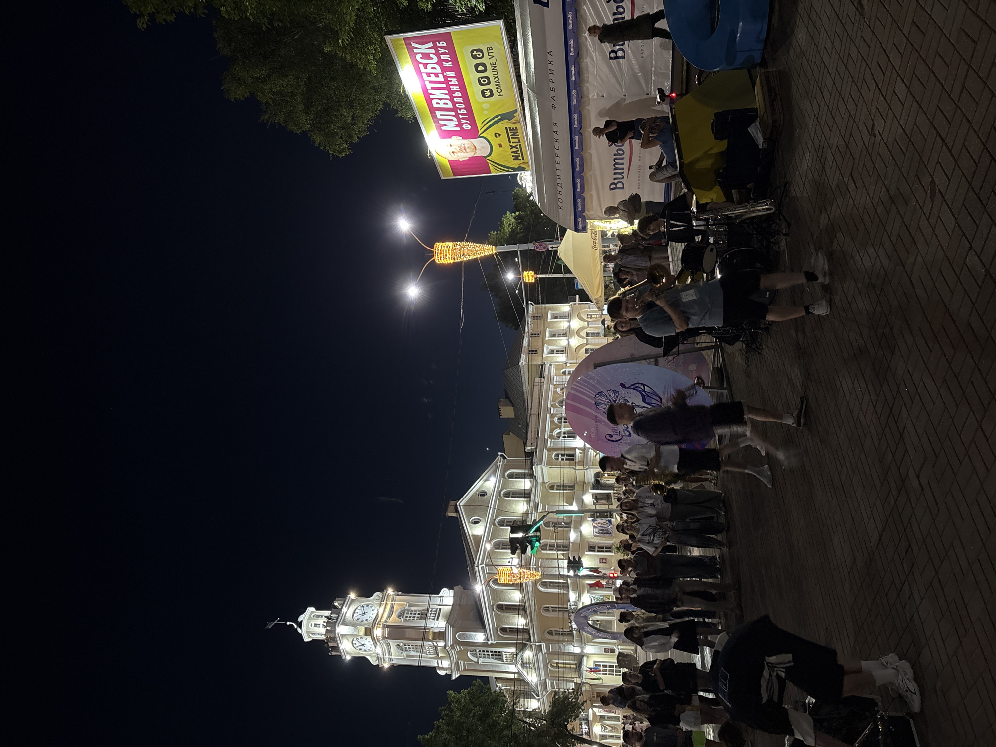 The image depicts a nighttime scene on a city street in Vitebsk, Belarus, likely in the vicinity of the Vitebsk City Hall (also known as the Governor's Palace). In the background, a prominent building with a clock tower and illuminated windows stands tall, characteristic of city hall architecture. In front of the building, a crowd of people is gathered on a paved area. Various elements suggest an event or festival: illuminated decorative lights resembling a giant caterpillar hang from a lamppost, and several large, colorful, circular structures are present, one with a graphic design.
A large banner on the right side advertises "FC MAXLINE VITEBSK FOOTBALL CLUB" with a portrait of a man in a football jersey. Below this, a white tent or stall displays the "BUMBA" logo repeatedly. In the foreground, a band is set up with a trombonist playing and a drummer behind him, accompanied by a saxophone. People are observing the musicians and milling about. The atmosphere appears lively, with the street illuminated by artificial lighting from the buildings, lampposts, and the event's decorations.
