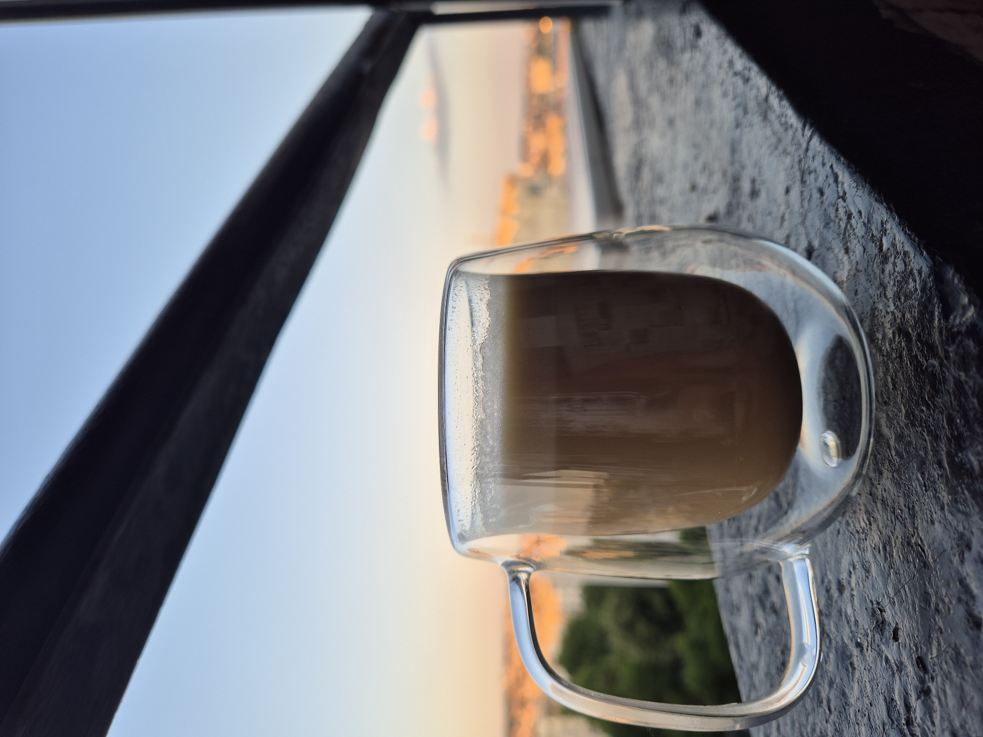 A double-walled glass mug filled with a frothy brown beverage sits on a textured dark surface, possibly a balcony railing, in the foreground. The mug is positioned slightly to the left of the frame, with its handle visible. Beyond the mug, a blurred city skyline is visible, illuminated by the warm hues of either sunrise or sunset. Lush green trees are also present in the mid-ground, adding a touch of nature to the urban landscape. The sky above transitions from a soft blue to a pale orange near the horizon, indicating the time of day. A dark, horizontal bar runs across the top of the frame, partially obscuring the sky.