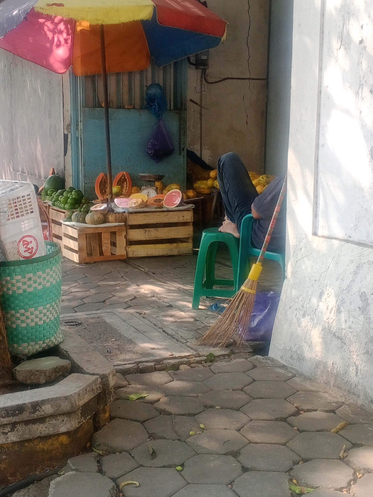 In a street-side market stall in Wonogiri, Indonesia, a fruit vendor sits on a green plastic stool, his legs crossed, wearing dark jeans and a dark t-shirt. He is partially obscured by a blue tarp and a broom with a yellow handle leans against his chair. The stall features an array of fruits, including papayas, grape-like green fruits, and what appear to be small pumpkins or gourds, displayed on wooden crates. Behind the fruit, a turquoise metal door is visible. An umbrella with segments of yellow, orange, pink, and blue provides shade over the stall. To the left, a woven green basket holds white plastic containers. The ground is paved with hexagonal stone tiles, and the scene is illuminated by sunlight, casting shadows on the pavement.