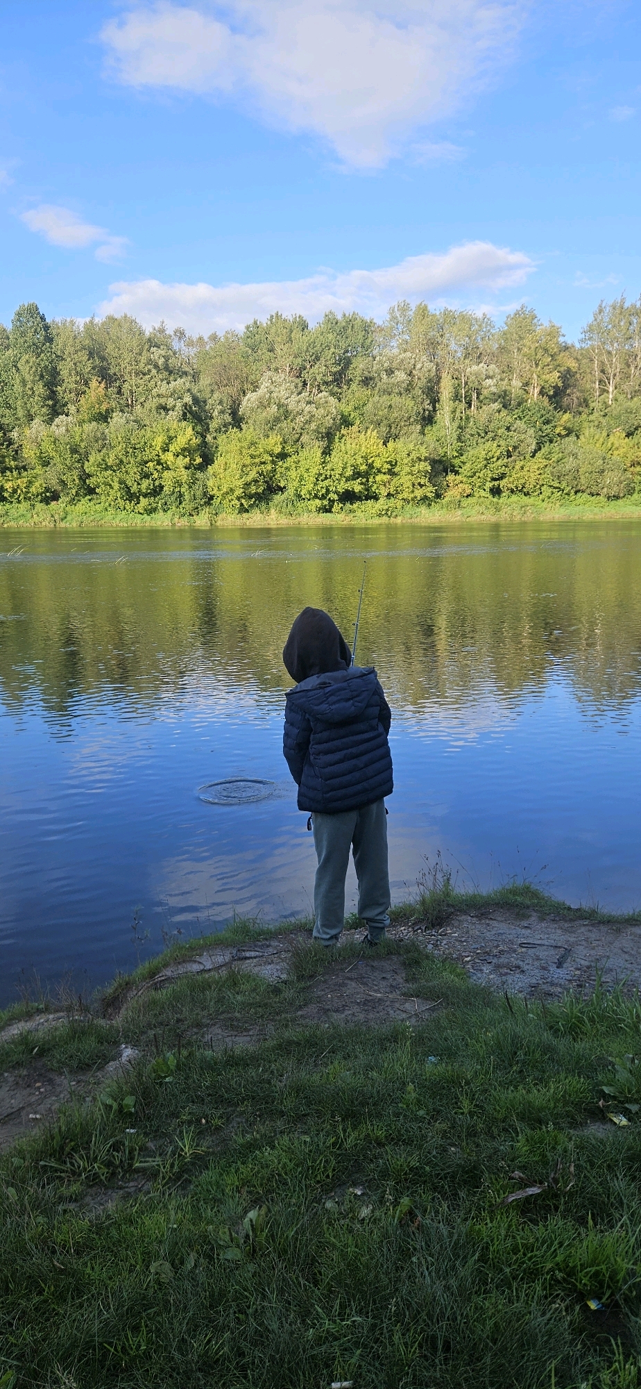 A person, seen from behind, stands on a grassy bank next to a wide river, holding a fishing rod. The person is wearing a dark hooded jacket and light-colored pants. The river is calm and reflects the trees on the opposite bank, which form a dense green treeline. The sky above is blue with scattered white clouds. The scene appears to be in a natural, unpopulated area.