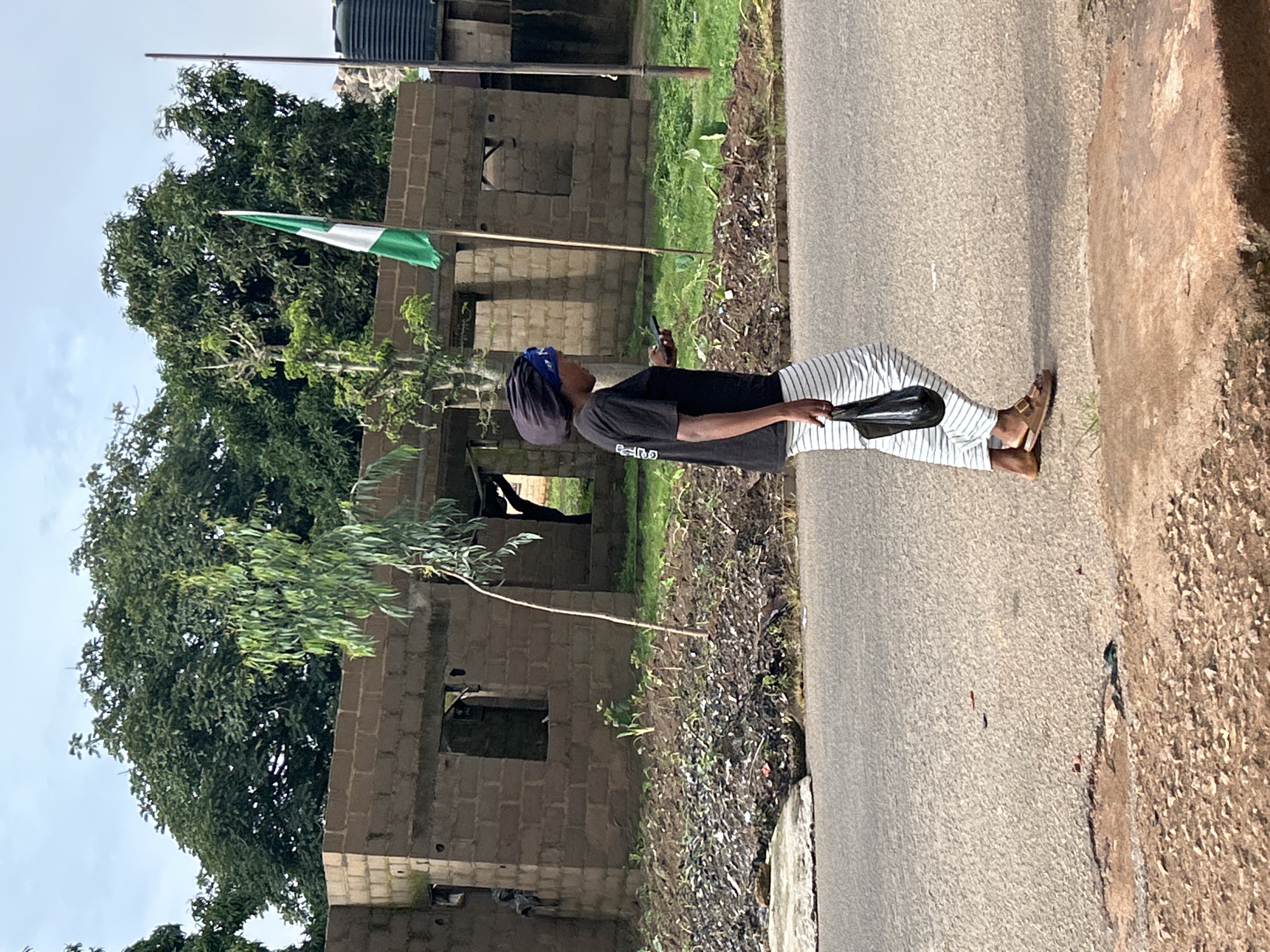 A medium shot captures a Black woman walking on a paved road in Jos, Nigeria. She is wearing a black t-shirt, a blue and black headwrap, white and black striped pants, and brown sandals. The woman is holding a black bag in her left hand and a dark smartphone in her right hand. To her right, a Nigerian flag waves on a pole. Behind her, a partially constructed building made of concrete blocks stands unfinished, with open doorways and windows. Lush green trees and vegetation surround the building. The ground in front of the building is covered in dirt and debris, with small plants growing in patches. The road surface is asphalt, with some cracks and patches visible. The sky is overcast with some patches of blue.