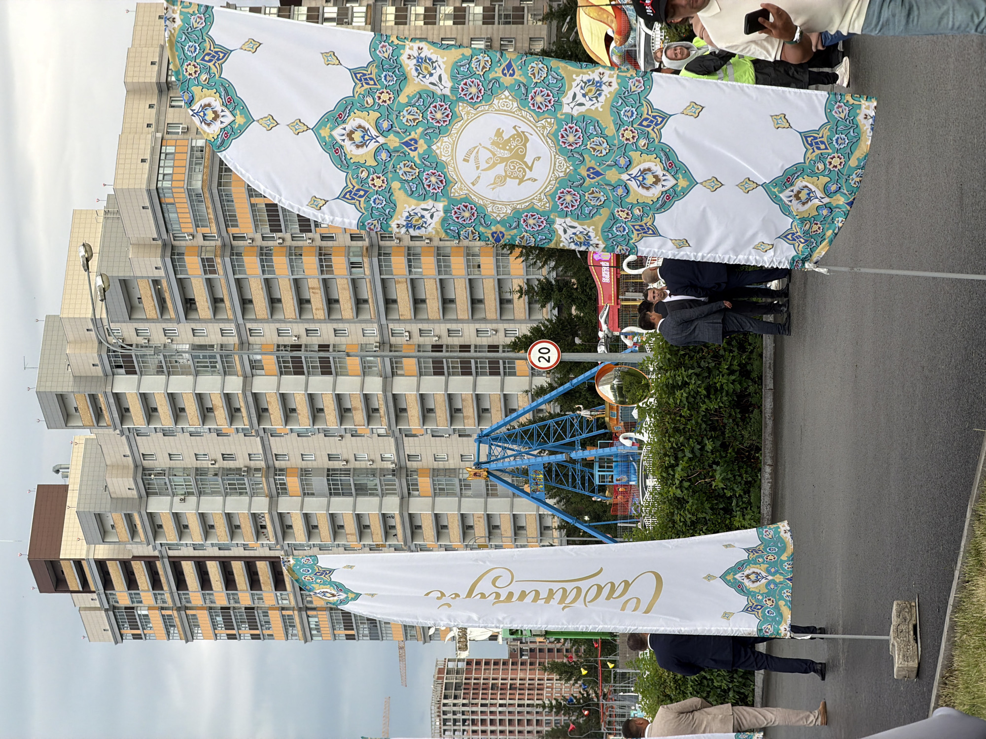 In an outdoor urban setting with a cloudy sky, two large white flags with ornate teal and gold patterns are prominently displayed. The flag on the left features gold calligraphy running vertically. In the background, a cluster of tall, modern apartment buildings with balconies and beige and grey facades dominates the skyline. To the right of the flags, a blue amusement park ride with a circular swinging mechanism is visible. Several people, dressed in formal attire and casual wear, are gathered near the flags and the amusement ride. The ground is paved, and a green hedge runs along the edge of the paved area. A traffic sign with the number "20" is visible on a pole. Construction cranes are also present in the distance, indicating ongoing development.
