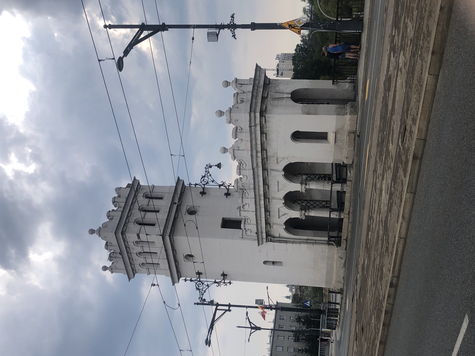 The image shows a white tower-like structure on a bridge in Warsaw, Poland, likely the Mokotowska Gate, part of the fortifications of the former Warsaw Citadel. The structure features arched openings on the ground level and decorative crenellations at the top. A street lamp with overhead wires is prominent in the foreground. Two individuals stand on the sidewalk near the tower, looking out. Tram tracks are visible on the road. In the background, other buildings and trees are visible, and the sky is overcast.