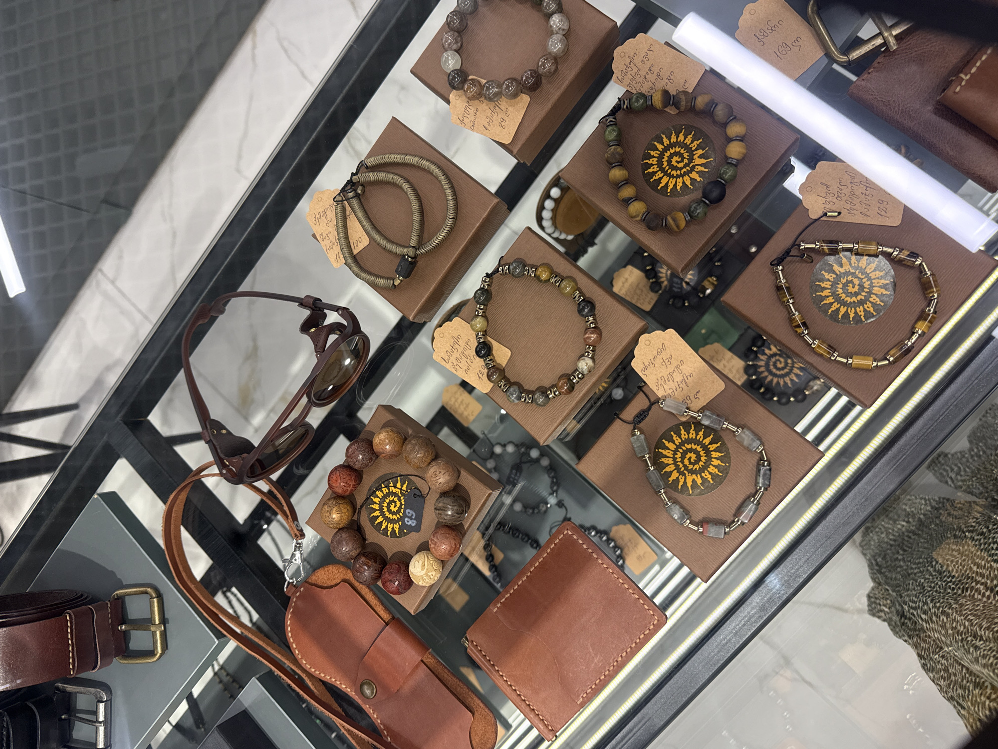 The image shows a glass display case filled with an assortment of handcrafted items, potentially from a shop in Batumi, Georgia, as indicated by the Georgian script on the price tags. The objects include several beaded bracelets displayed on small brown boxes, a pair of brown sunglasses, a leather belt with a brass buckle, a leather wallet, and a leather case, possibly for a phone or small accessory. The bracelets have different bead patterns, and each is placed on a box featuring a sun-like logo. Price tags written in Georgian are attached to some of the items. The general atmosphere suggests a boutique or market stall offering handmade leather goods and jewelry.