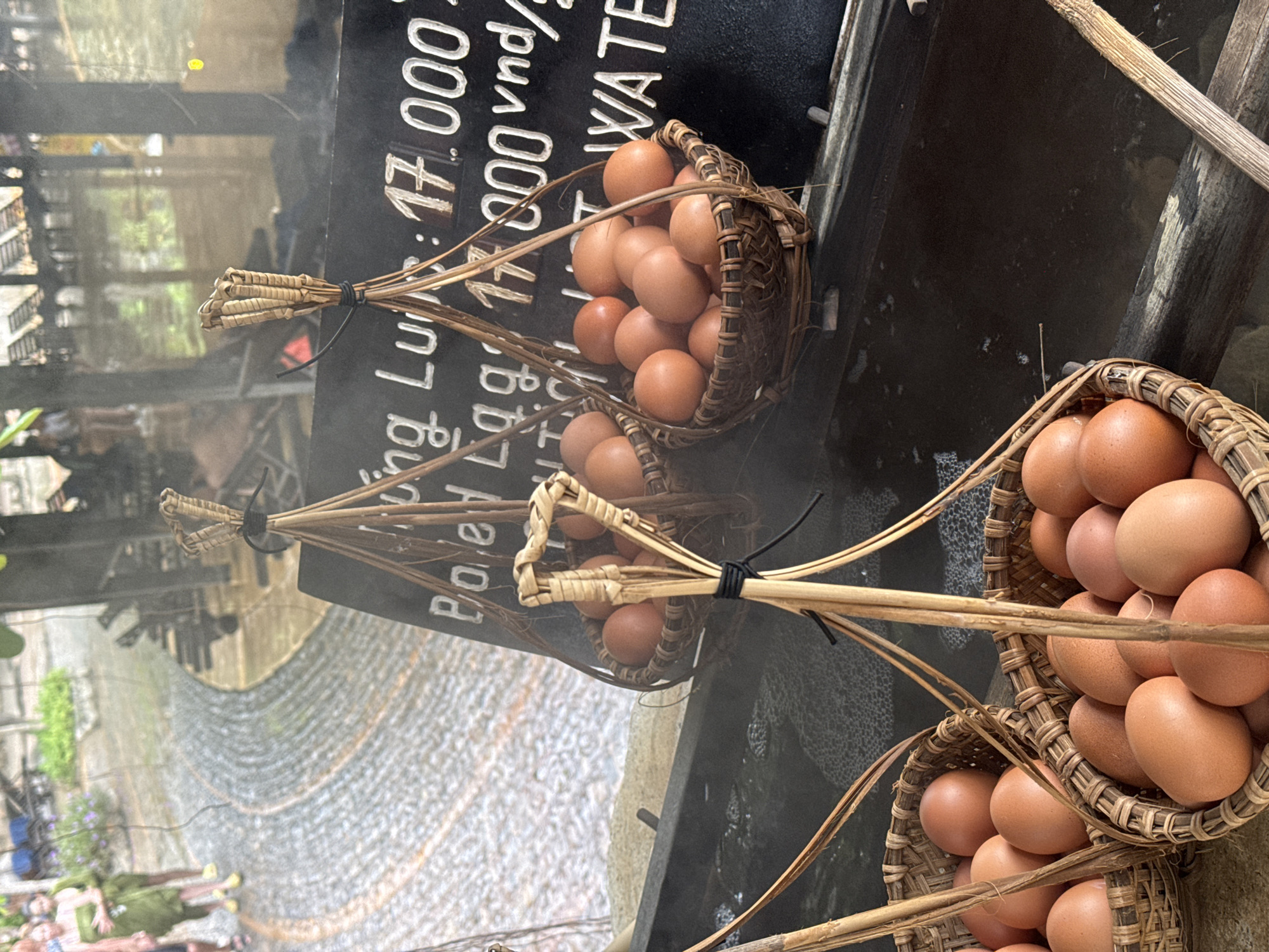 In Nha Trang, Vietnam, this image depicts a hot spring facility. In the foreground, several woven baskets filled with brown eggs are suspended over a steaming body of water. The eggs appear to be raw and are the primary subject of the image. In the background, a person in a green sarong is visible, suggesting the presence of other patrons or staff. A sign with text, partially obscured, indicates pricing in Vietnamese Dong. The overall environment suggests a place where eggs are boiled in natural hot springs.