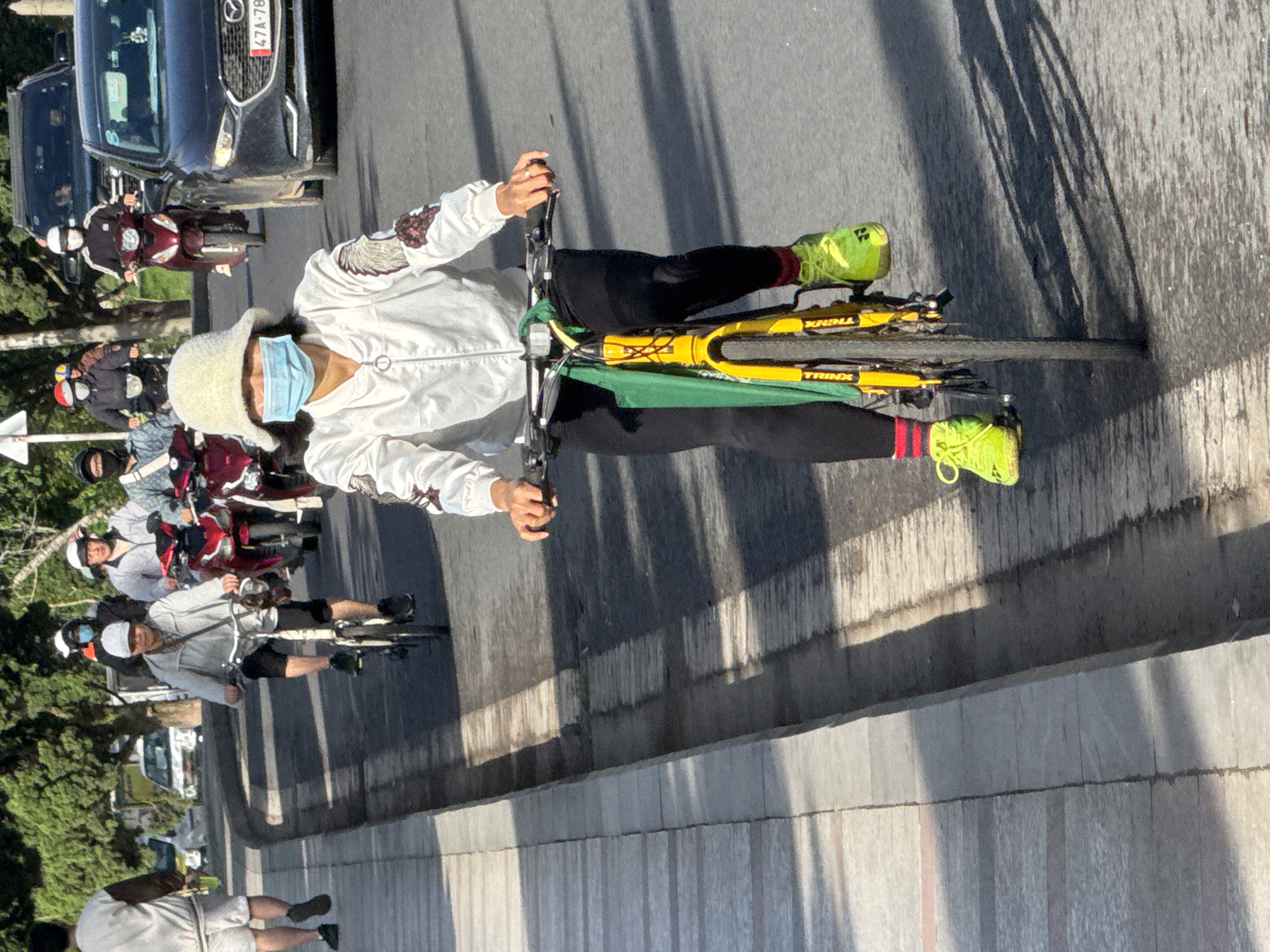 In Lam Vien - Da Lat, Vietnam, a person wearing a white bucket hat and a light-colored bomber jacket with a blue face mask is riding a yellow bicycle with a green cloth draped over the handlebars. They are wearing black leggings and neon yellow sneakers with red socks. The bicycle has a silver frame and black tires, and the spokes are visible. The cyclist's hands are on the handlebars, and their body is leaning slightly forward. The bicycle is on a paved road with a grey sidewalk to the left. In the background, other people are riding motorcycles. One person is on a red motorcycle, and another is on a dark-colored motorcycle. A black car is also visible behind the cyclists. The scene is illuminated by sunlight, casting shadows on the road.