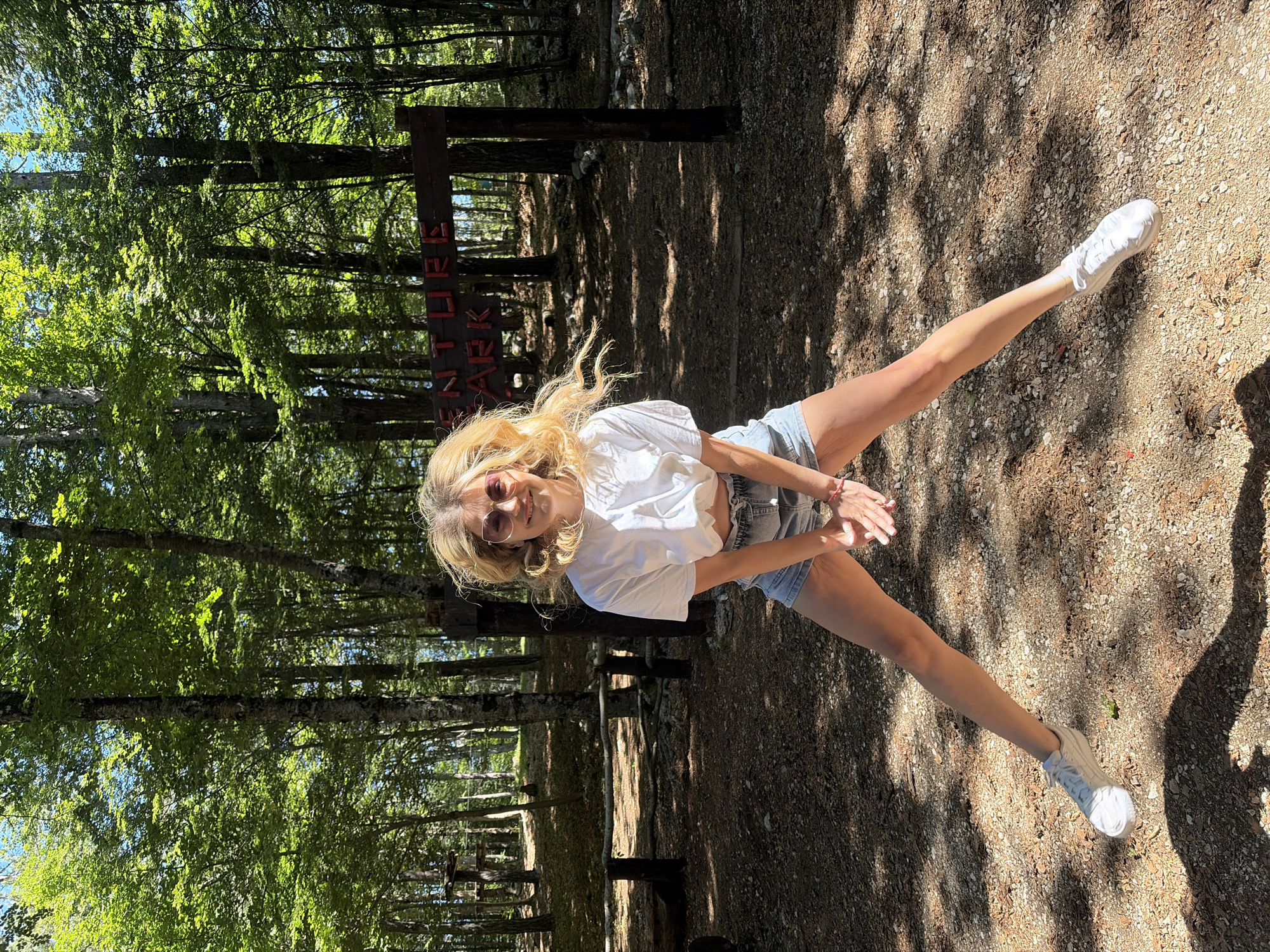 The image depicts an outdoor scene in a forested area, identified as Mali Bostur, Montenegro. The central subject is an adult female captured mid-air in a jumping motion. She has light-colored hair, which appears disheveled from movement, and wears large-framed sunglasses with a reddish tint. Her attire consists of a white short-sleeved cropped t-shirt and light blue denim shorts. Her hands are clasped at waist height, and her legs are spread wide with her feet elevated off the ground, clad in white athletic shoes.
In the background, a wooden sign with red lettering is visible, partially reading "ENTURE PARK," indicating an adventure park setting. Numerous deciduous trees with green foliage fill the background, creating a dense forest environment. Sunlight filters through the tree canopy, casting dappled light and shadows across the ground. The ground surface is composed of dry earth, small pebbles, and scattered organic debris. To the left of the frame, subtle wooden structures, consistent with an adventure park's course elements, are visible among the trees, alongside a low wooden barrier.