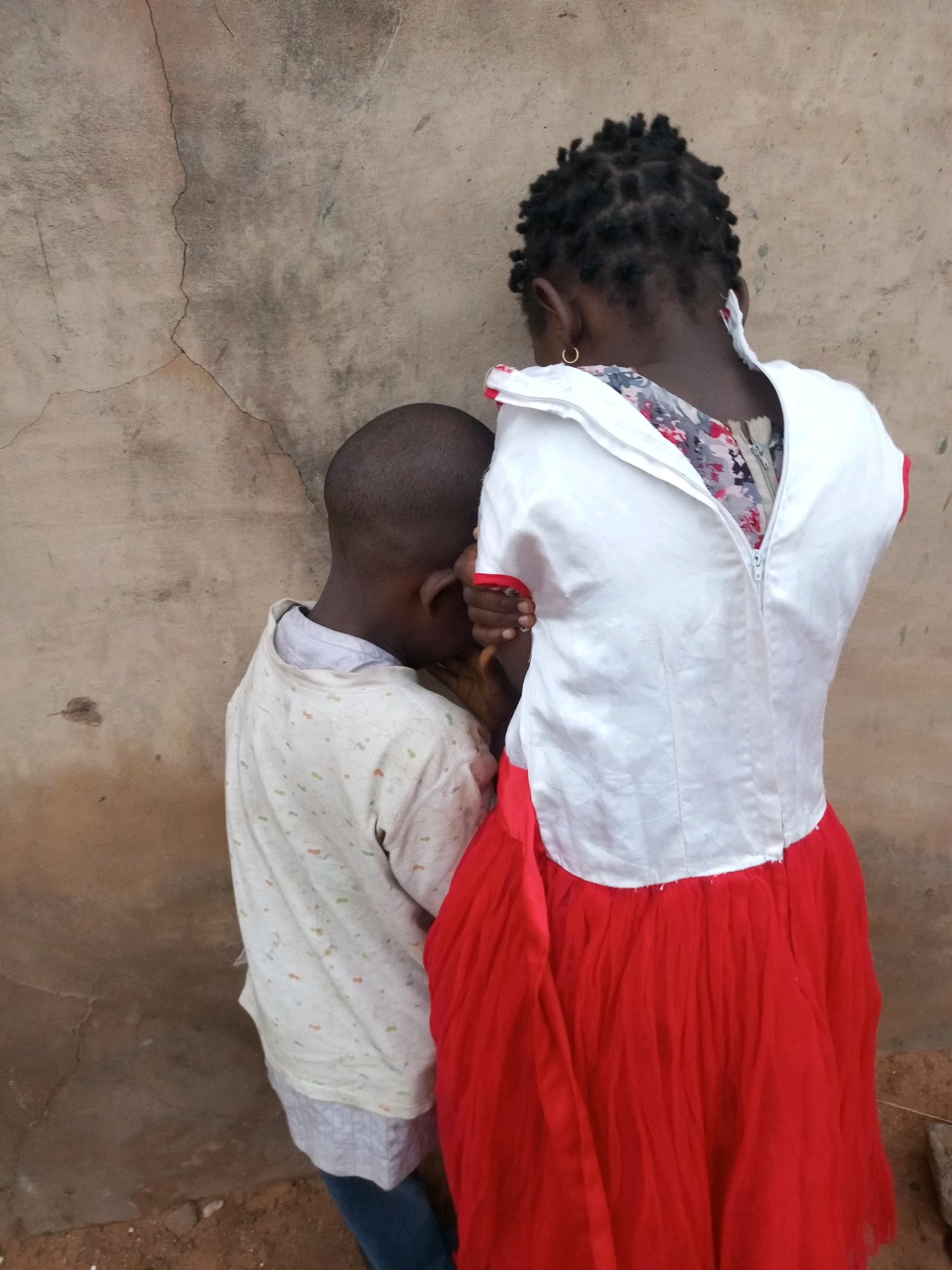 In Tempure, Nigeria, two children stand with their backs to the camera, leaning against a rough, cracked concrete wall. The child on the left, a boy with a shaved head, wears a light-colored shirt with a subtle patterned design. He is positioned slightly in front of the other child and appears to be looking down, with his head bowed and his arm resting near his face. The child on the right, a girl, is wearing a white bodice with a red tulle skirt. The bodice is zippered at the back, revealing a floral patterned fabric underneath. She has dark, braided hair and is wearing a small hoop earring in her visible ear. Her left hand is visible, touching the boy's shoulder or neck, suggesting an interaction or moment of comfort between them. The ground beneath them is dirt and gravel.