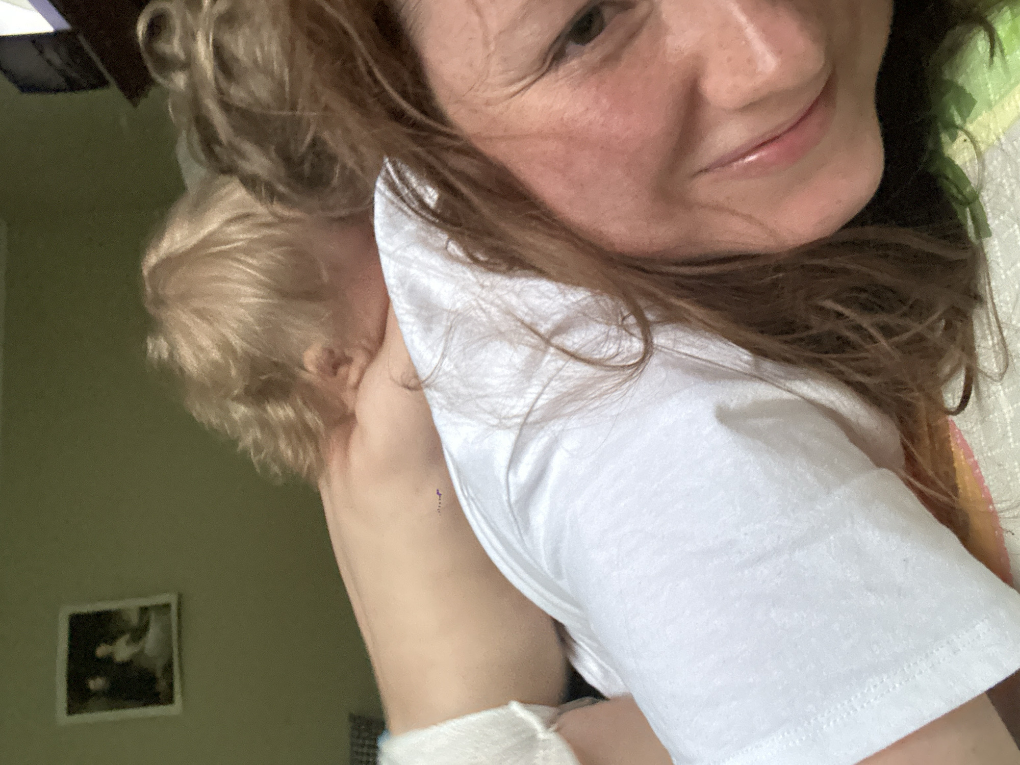 A woman is holding a young child with blond hair. The woman is wearing a white t-shirt and smiling at the camera. The child is turned away from the camera, with their head resting on the woman's shoulder. In the background, there is a green wall with a framed picture hanging on it. The picture appears to be of a child. A portion of bedding is visible in the top right corner. The city of Nizhny Novgorod, Russia is not visibly present in the image.