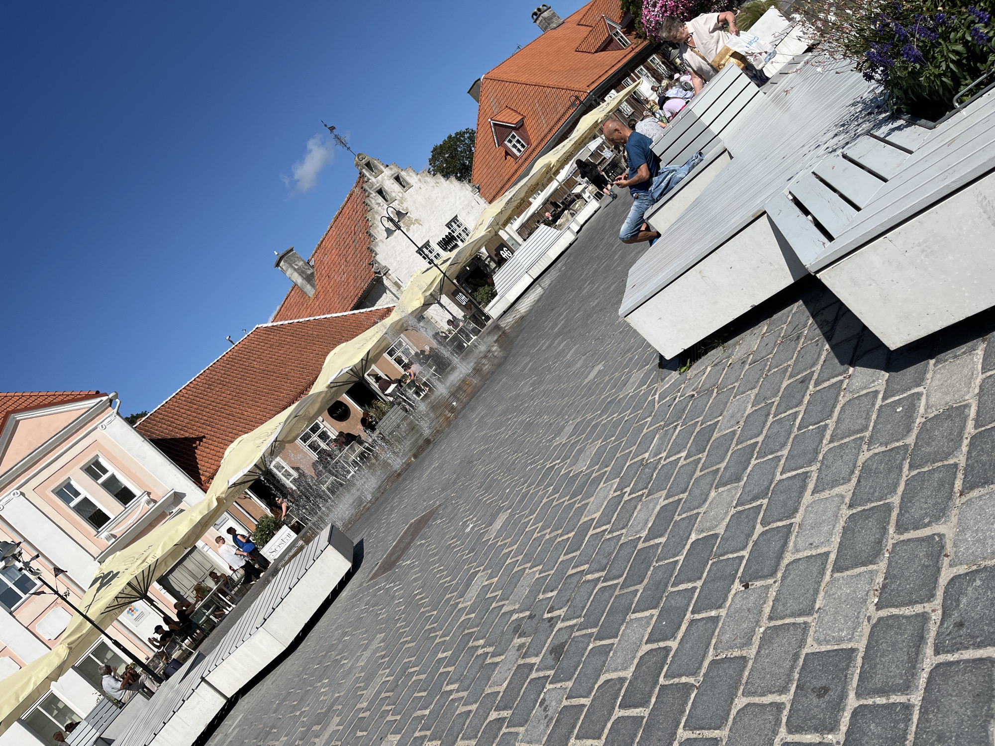 In Kuressaare, Estonia, a sun-drenched public square is paved with cobblestones. A man, seated on a modern, slatted bench, is engrossed in his phone. Adjacent to him, another person is also seated on the bench, facing away from the camera. In the background, an outdoor cafe features patrons seated at tables beneath yellow umbrellas, with a fountain providing a refreshing spray. Buildings with red-tiled roofs and light-colored facades line the square, one featuring a distinctive turret. The sky is a clear, bright blue, indicating a pleasant day.