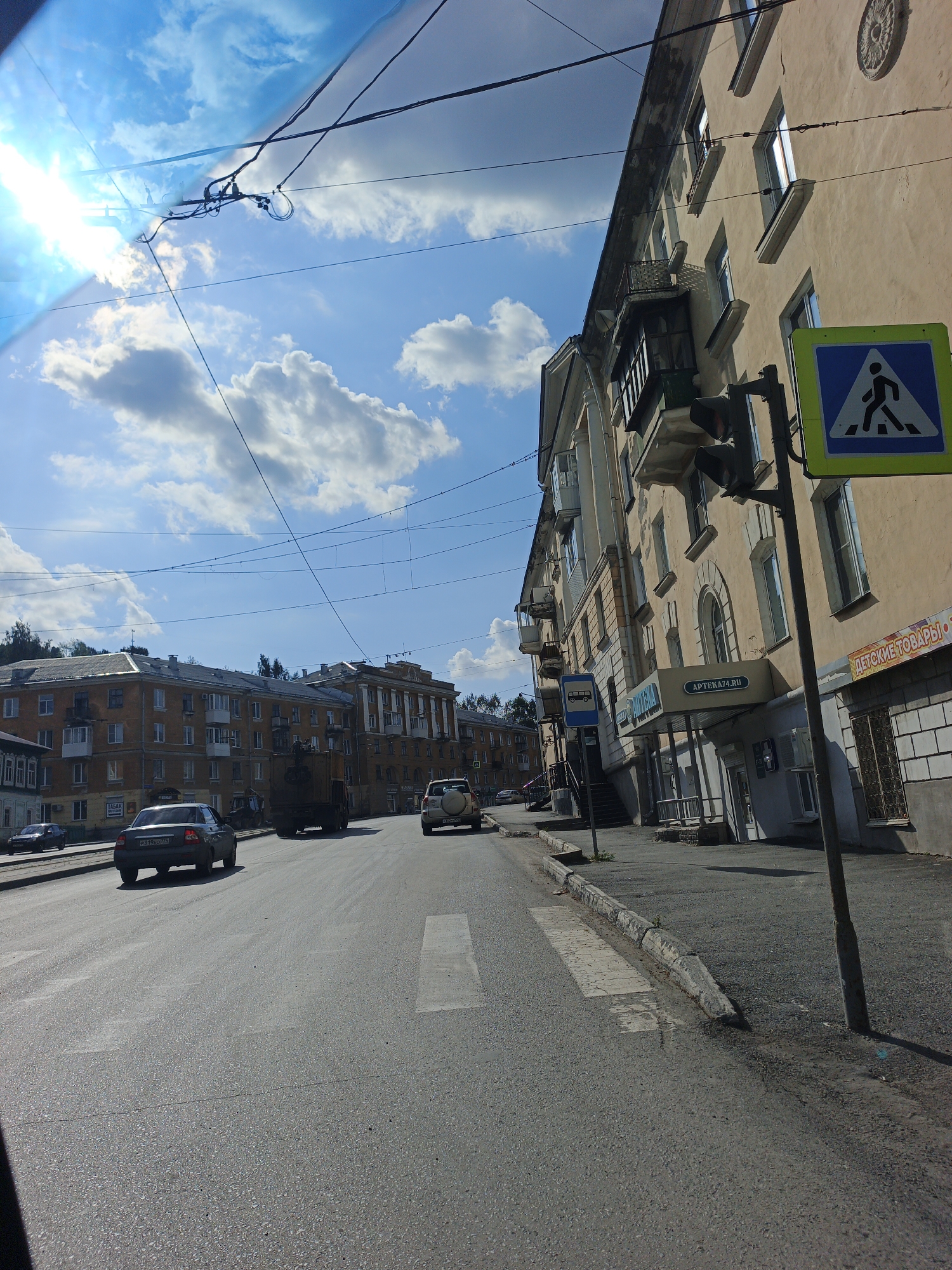 The image depicts a street scene in Zlatoust, Russia, on a sunny day with scattered clouds. A pedestrian crossing is visible in the foreground on the right side of the road, marked by white stripes. A blue and white pedestrian crossing sign is mounted on a pole next to it. A traffic light is also present on the same pole.
Several buildings line the street. On the right, a multi-story building with light-colored facade features multiple windows and balconies. Signs visible on this building include "APTEKA.RU" and "ДЕТСКИЕ ТОВАРЫ" (Children's Goods). On the left side of the street, more multi-story buildings with yellow facades are visible.
Traffic on the road includes a dark-colored car in the left lane, a dark truck in the right lane, and a light-colored SUV in the far right lane. Overhead power lines crisscross the sky, and the sun is bright, causing lens flare in the upper left portion of the image. The road surface is asphalt.