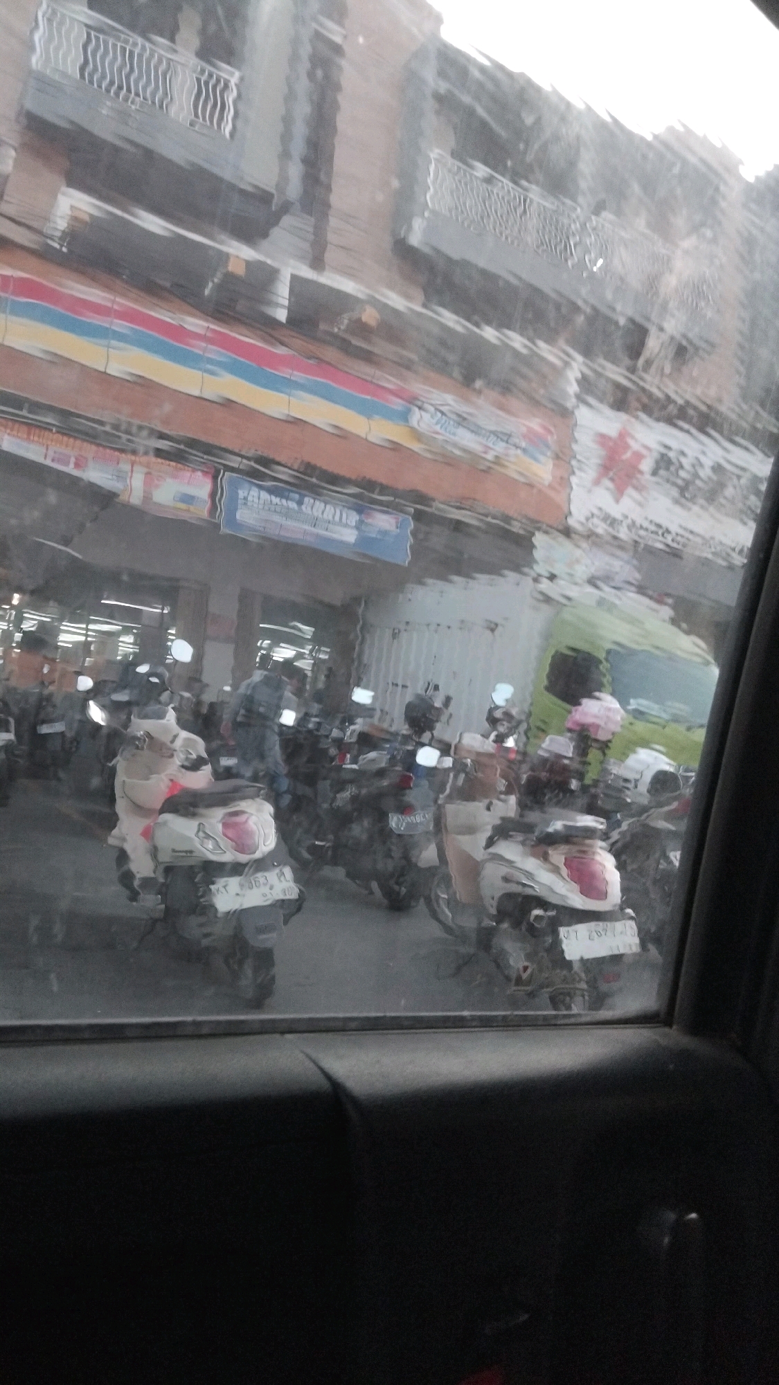 The image, taken from inside a vehicle, shows a street scene in Samarinda, Indonesia, possibly in a commercial or mixed-use area with a building in the background that appears to be a multi-story structure with balconies. A striped awning or sign with red, yellow, and blue bands is visible on the building. Below this, there is a blue sign that reads "PARKIR GRATIS" (Free Parking). The foreground is dominated by a large number of parked motorcycles. Some of the motorcycles are white, and one has a license plate that appears to read "KT 6063 KL". A person in a gray shirt is standing among the motorcycles. To the right, a green truck is partially visible. The overall scene suggests a busy area with a parking lot for motorcycles. The image quality is somewhat obscured, likely due to the view through a window with water droplets or smudges.