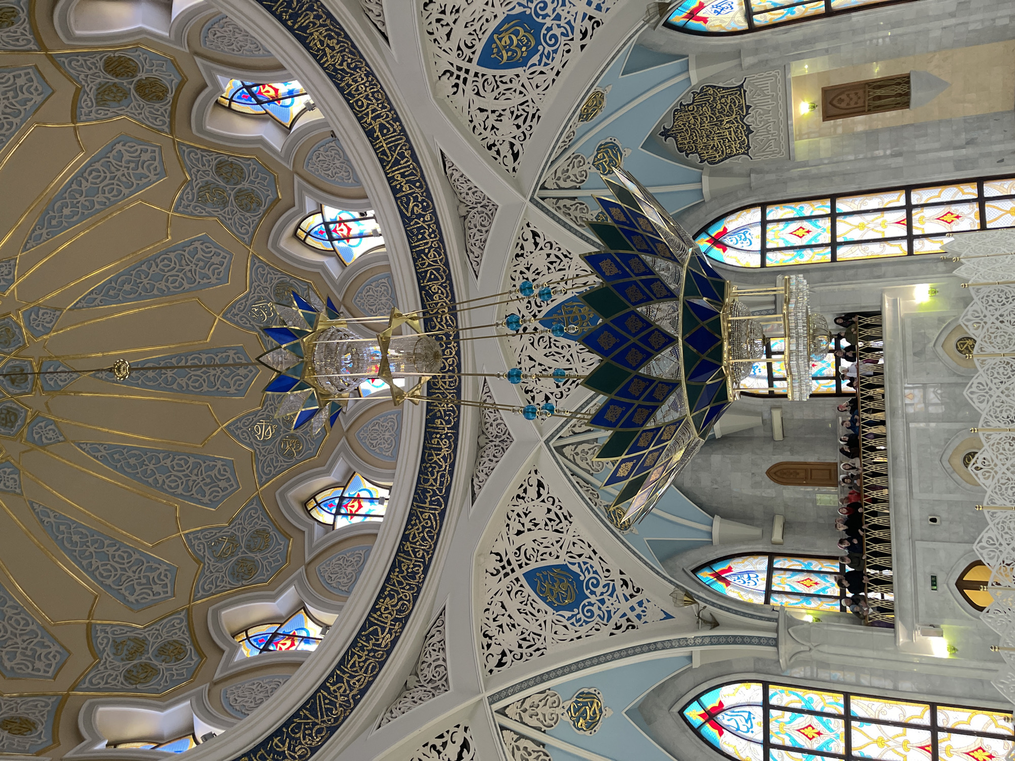 The image shows the interior of the Kul Sharif Mosque in Kazan, Russia. The camera is pointed upward, capturing the ornate ceiling. The ceiling is a mix of geometric patterns, Arabic calligraphy, and stained-glass windows. There is a large chandelier hanging from the center. A balcony filled with people in the background.