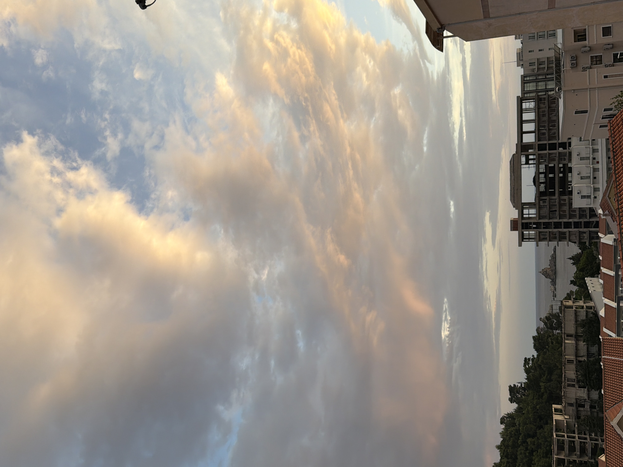 The image shows an urban landscape with a view towards the sea, likely taken from an elevated position in Petrovac na Moru, Montenegro. The foreground is dominated by a sky filled with clouds, some catching the golden light of either sunrise or sunset. Below, various buildings are visible, including multi-story residential or commercial structures. Some buildings appear to be under construction or in a state of partial disrepair. In the distance, the sea horizon is visible, with a small rocky islet emerging from the water. A dense patch of trees or forest separates the buildings from the shoreline. No people or animals are visible in the image.