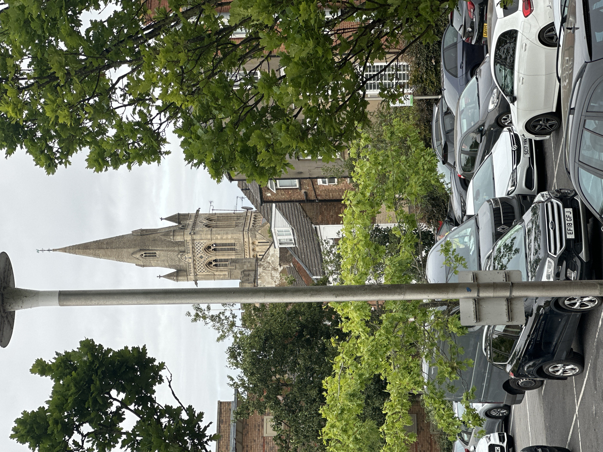 The image shows a view of a car park with several vehicles parked. In the background, a church tower with a spire is visible, surrounded by trees and buildings. The sky is overcast. A lamppost is prominently featured in the foreground, partially obscuring the view of the cars and the church. One of the cars in the foreground is a black SUV with license plate EF18 GFJ. Other cars include a grey van and several passenger cars of various colors. The overall setting appears to be an urban environment. The church in the background is St. Andrew's Church, located in Rugby, United Kingdom.