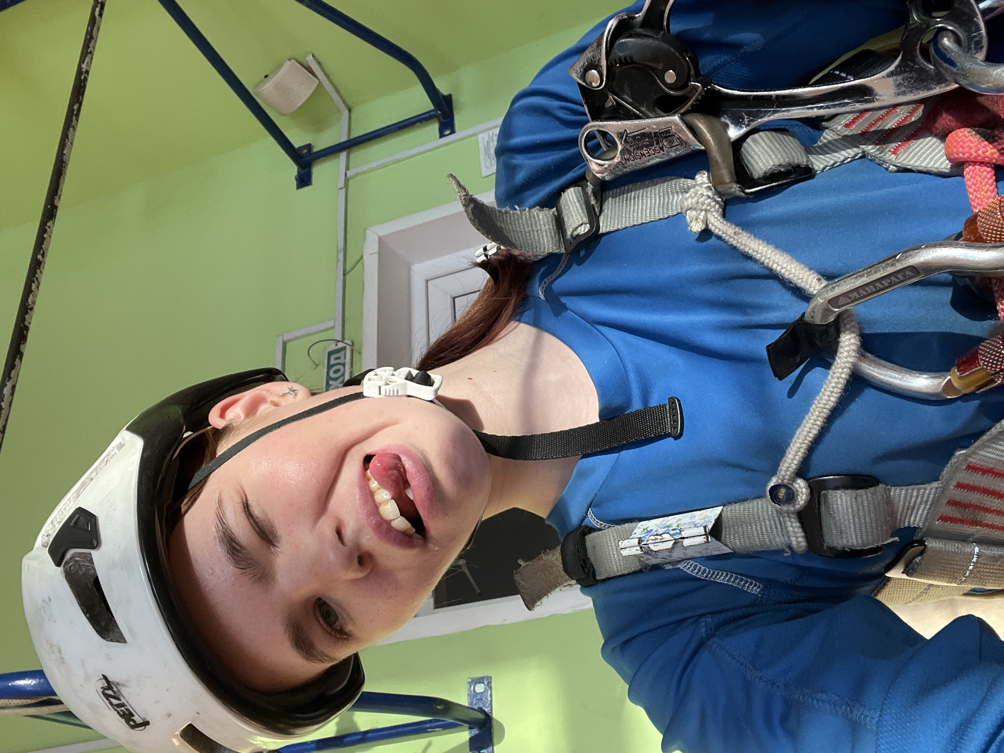 A young woman wearing a white climbing helmet and a blue climbing harness is the primary subject of the image. She is looking at the camera, sticking her tongue out playfully. The helmet is a Petzl brand. The background is a light green wall with blue and white fixtures. The location is a climbing facility in Chaikovsky, Russia, as indicated by the harness markings.