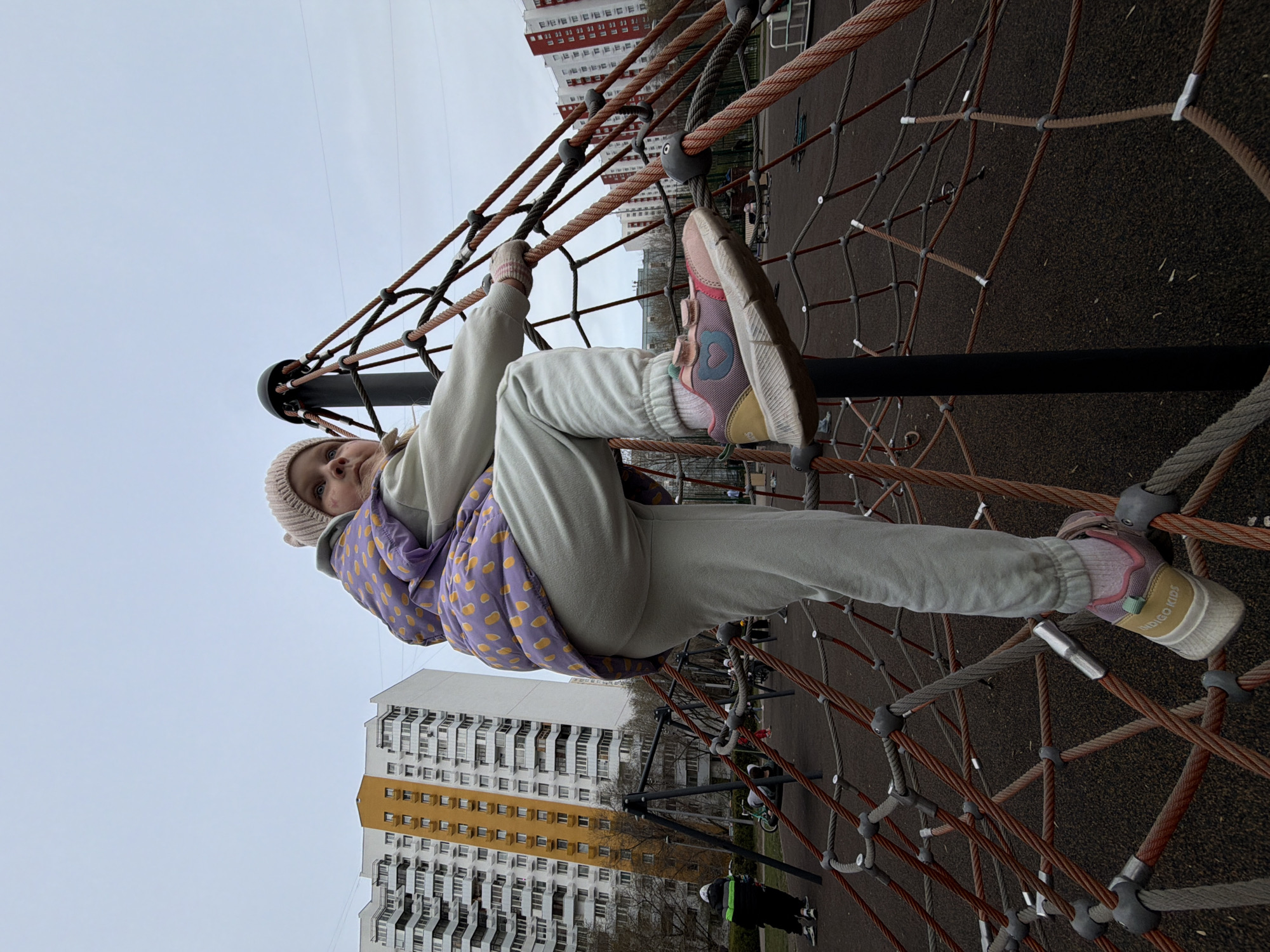 A young girl, likely a child or pre-teen, is captured actively climbing or posing on a large rope pyramid structure at an outdoor playground in Moscow, Russia. She is positioned with one leg bent, her foot resting on a rope, and both hands gripping ropes, indicating engagement with the climbing net. She looks directly at the camera with a neutral to slightly curious expression.
The girl is dressed in a light grey tracksuit, a purple puffy vest adorned with yellow polka dots, and a light pink beanie. Her colorful sneakers feature shades of pink, yellow, blue, white, and grey, and the text "INDIGO KIDS" is visible on the sole of her right shoe.
The playground features a robust climbing net constructed from thick orange and grey ropes connected by black plastic or metal joints and central support poles. The ground beneath the structure is a dark, likely rubberized safety surface. In the background, several tall, multi-story apartment buildings characterize the urban residential setting. These buildings are mainly white and grey, with one featuring red upper floors and another displaying yellow panels on its lower section. A small portion of another person, wearing a dark jacket with a green stripe and a helmet, and a bicycle are faintly visible near the base of the buildings. The sky is overcast and grey, suggesting a cloudy day with cool or mild weather. The photograph is taken from a low angle, looking upwards towards the girl and the climbing structure.