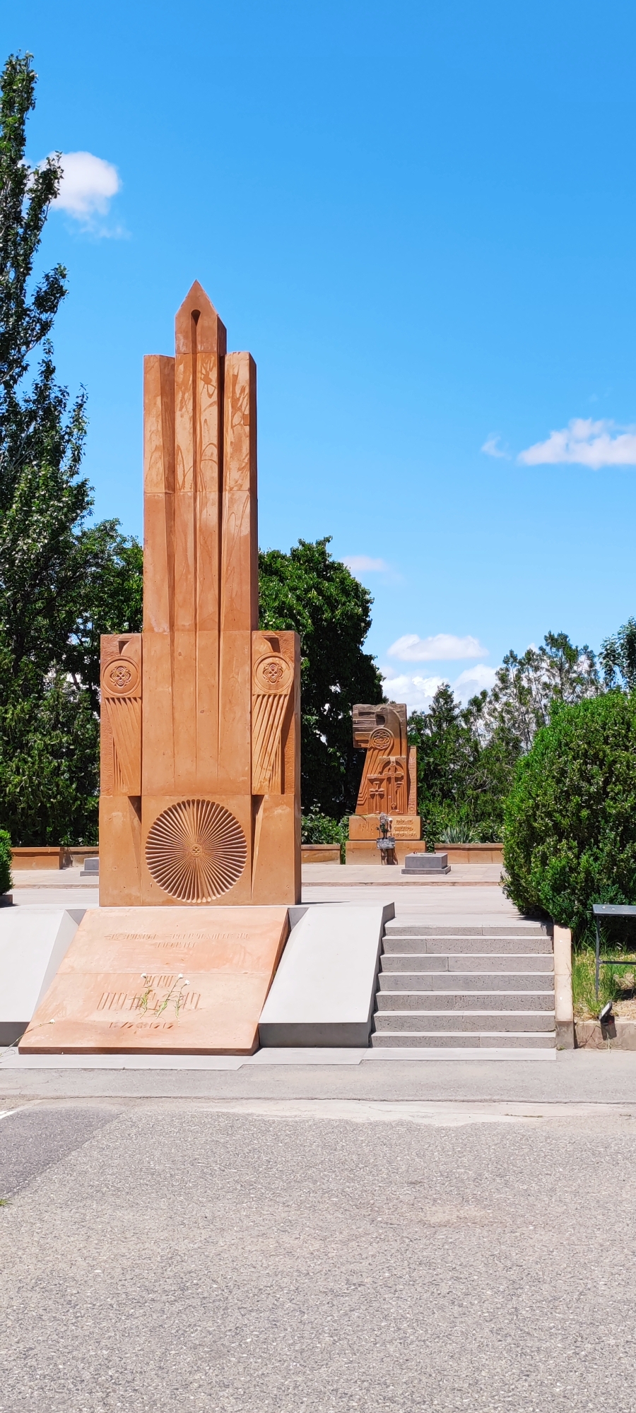 The image depicts an outdoor scene with a monument in the foreground, likely in the city of Araks, Armenia. The monument is constructed from reddish-orange stone and features intricate carvings. The main structure is tall and slender, with a pointed top. To its left, there's a wider base with a circular, sunburst-like carving. Inscribed on a slanted lower section of the monument are lines of text and what appear to be sprigs of foliage.
In the background, several trees with lush green foliage are visible, creating a natural backdrop against a clear blue sky with a few scattered white clouds. To the right of the main monument, another, smaller monument made of similar stone is partially visible. This secondary monument also features carved elements, including a cross-like symbol. A set of concrete stairs leads up to a higher level in front of the monument. The ground in the foreground is paved with asphalt. The overall impression is one of a memorial site or public park.