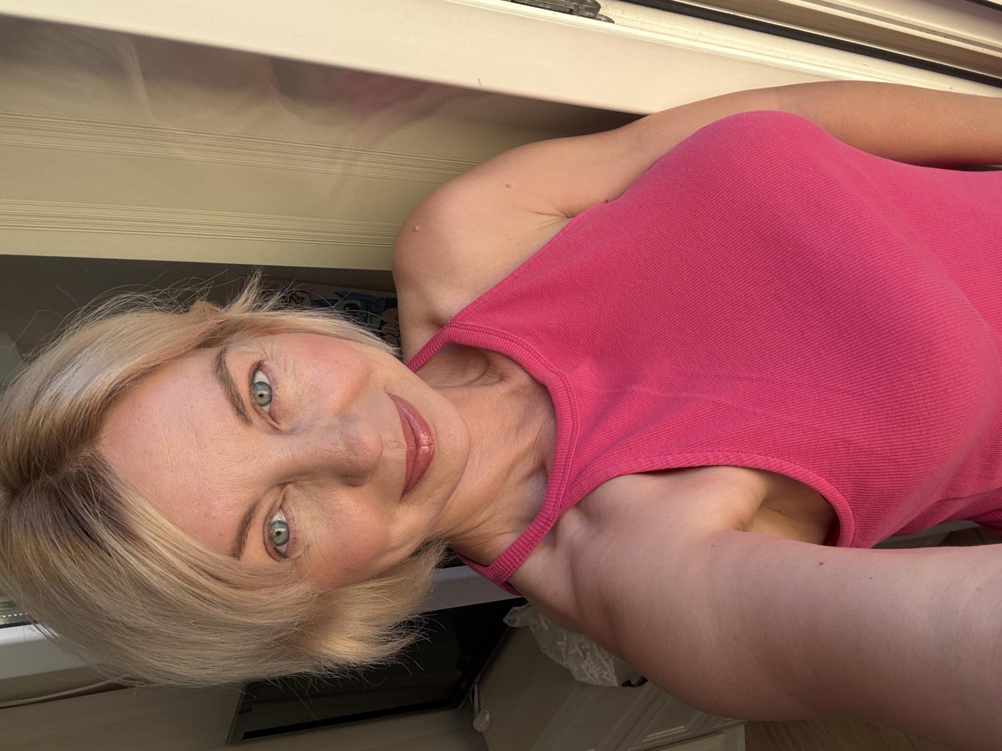 A blonde woman with blue eyes and pink lipstick looks directly at the camera. She is wearing a bright pink tank top with a halter neckline. Her hair is short and styled around her face. The background appears to be an indoor setting with neutral-colored walls and trim, possibly a doorway or closet. There is a hint of a dresser or furniture to the left of the frame. The lighting suggests natural light coming from the right. There are no specific landmarks or indicators that would place this image in Sochi, Russia.