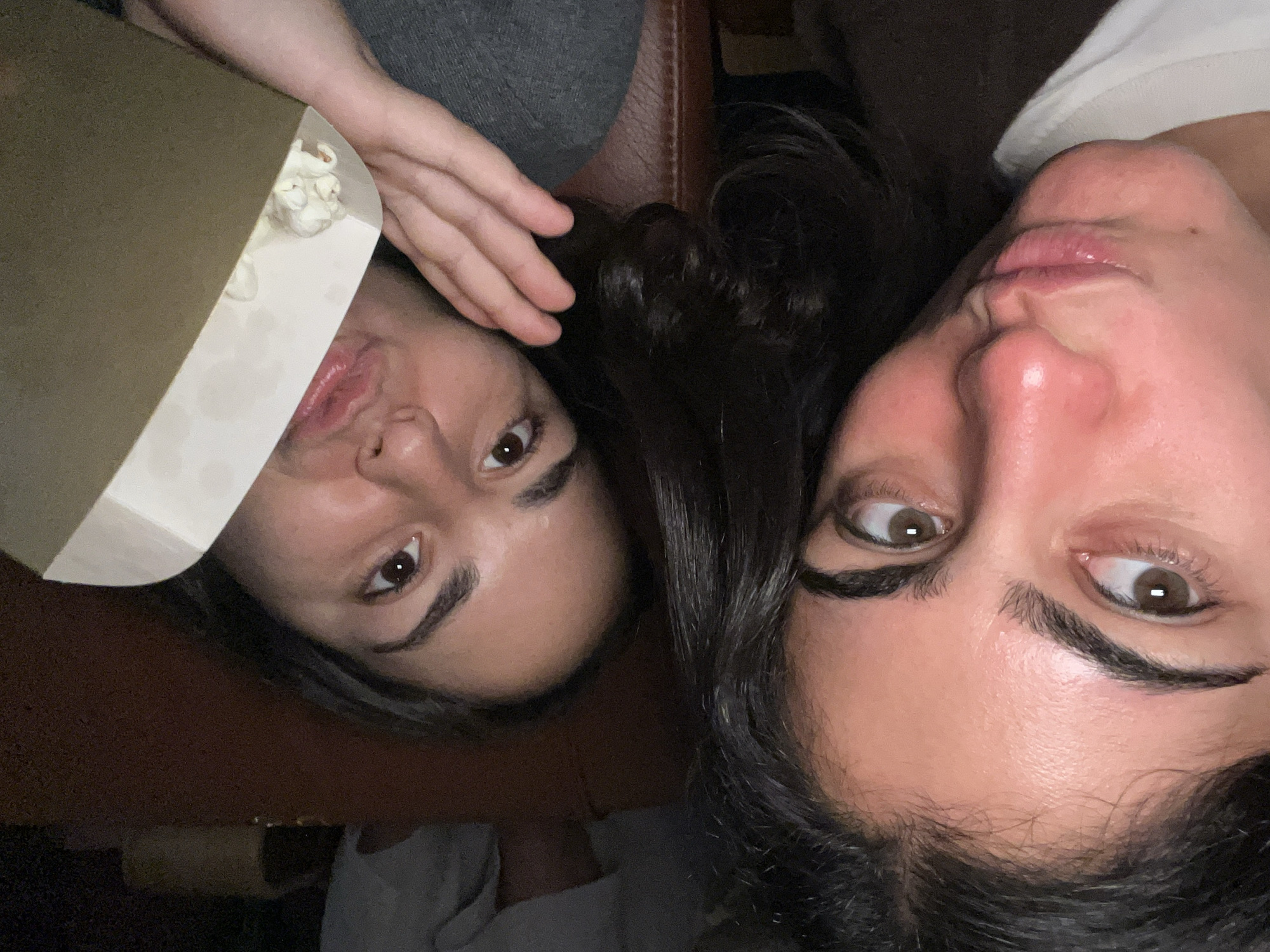 Two women are lying close together, their faces angled towards the camera. One woman holds a partially opened carton containing white contents, possibly popcorn or a similar snack food. The environment appears to be indoors, possibly on a leather couch or similar surface. The specific location in Paris, France is indiscernible from the image. No specific actions or interactions are evident beyond the posing for the photograph.