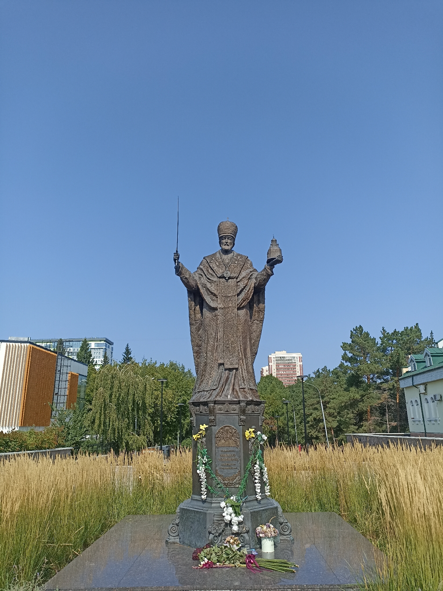 The image shows a bronze statue of a robed figure wearing a mitre and holding a sword in its right hand and a small cross atop a globe in its left. The statue is positioned centrally in the frame, on a dark granite pedestal adorned with flowers. The background consists of a clear blue sky, tall dry grass, trees, and modern buildings. The statue is located in the city of Novosibirsk, Russia, specifically in front of the Novosibirsk State Opera and Ballet Theatre.