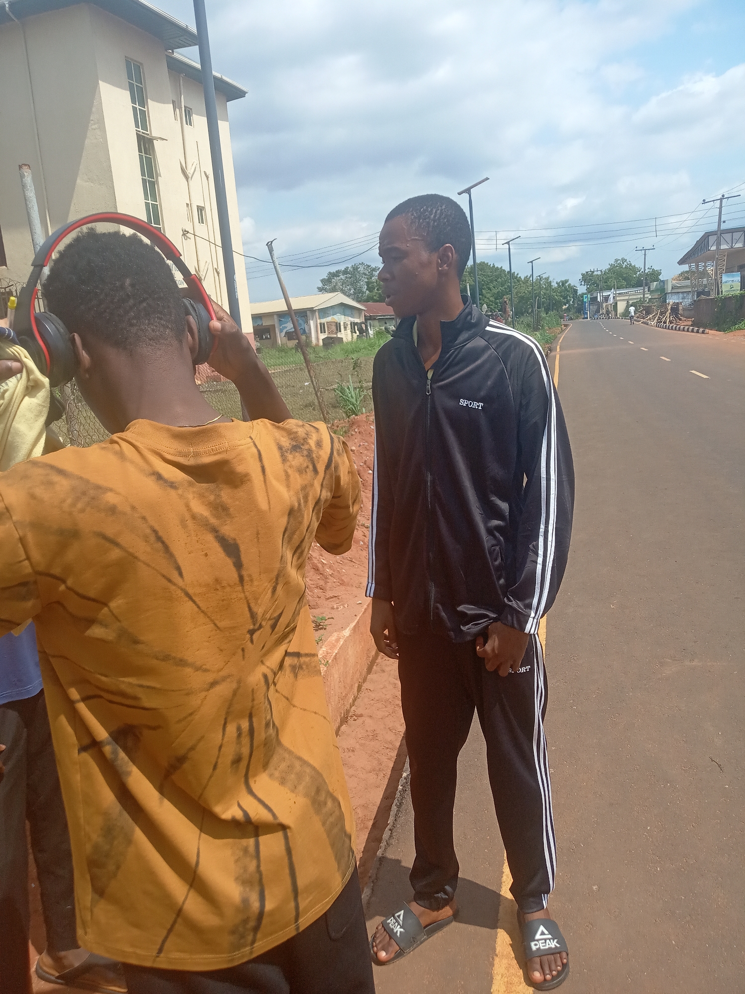 The image depicts two young men outdoors on a street or roadside, likely in Nsugbe, Nigeria, under a partly cloudy sky during daytime.
In the foreground, with his back to the camera, is a man wearing a mustard yellow t-shirt with a dark, tie-dye or distressed pattern. He has short, dark hair and is holding black headphones with red accents to his ears, appearing to adjust them.
To his right, in the middle ground, stands another man looking to the left with a focused or pensive expression. He is dressed in a black tracksuit, consisting of a zipped jacket and matching pants, both adorned with white stripes running down the sides of the sleeves and legs. The jacket has "SPORT" written in white on the left chest, and the pants also show "SPORT" text on the thigh. He wears black slide sandals, each featuring a white "PEAK" logo and a triangular design.
The background shows an asphalt road extending into the distance, flanked by a concrete curb and reddish soil. On the left, a tall, light-colored building with prominent vertical windows is partially visible, behind a metal pole and a fence. A portion of a sign on this building displays the text "NATION." Further back, utility poles, power lines, modern streetlights, other buildings, and green trees dot the landscape. The scene conveys a casual, everyday outdoor setting.