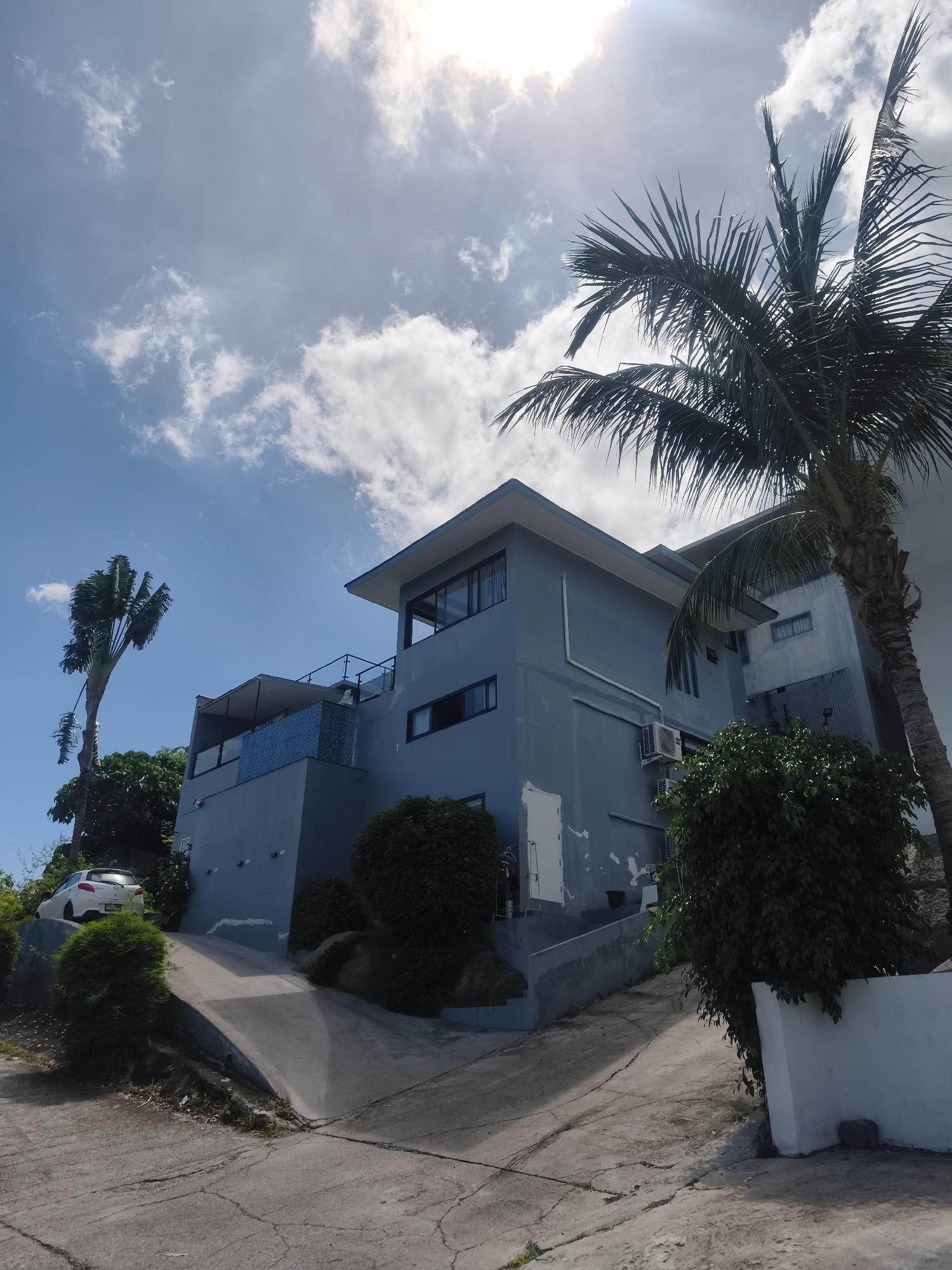 The image depicts a multi-story blue building with a flat roof, possibly a residence or hotel, situated on a slope. A paved driveway curves up towards the building. Several palm trees are visible, one large one prominently in the foreground on the right, and another thinner one on the left. The building features balconies, windows, and exterior air conditioning units. Some greenery and bushes are planted around the building and along the driveway. A white car is parked on the left side of the driveway. The sky is blue with scattered clouds, and sunlight is visible. The scene suggests a tropical or subtropical environment. The location is identified as Ko Samui, Thailand.