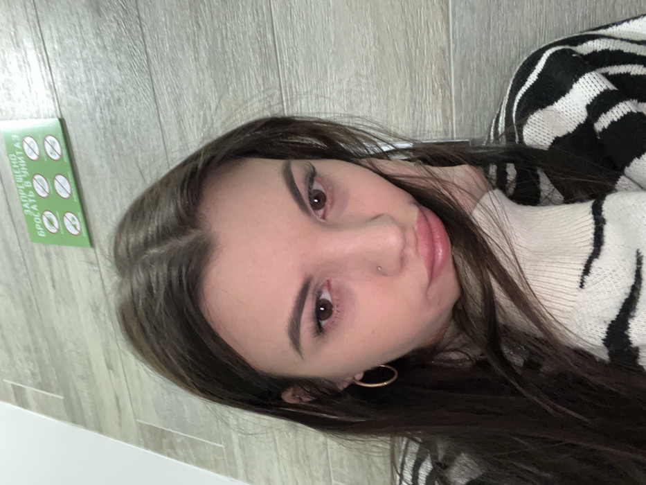 A woman with long, dark hair and light skin looks at the camera with brown eyes. She has a nose piercing and is wearing gold hoop earrings. She is wearing a zebra-striped sweater with a white turtleneck. A green sign with icons and text in Russian is visible in the background. The background appears to be a gray-toned wooden floor.