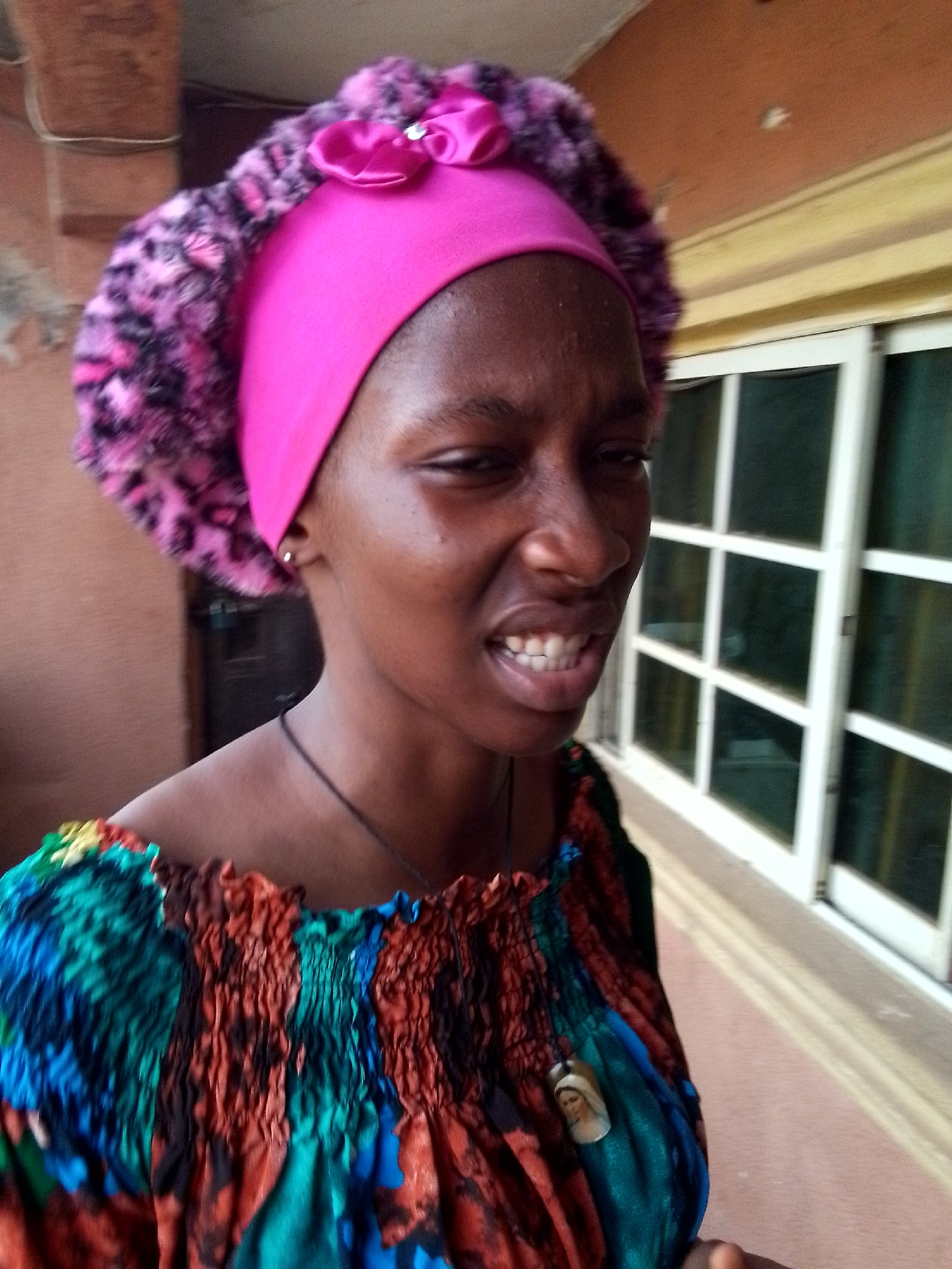 A dark-skinned woman with short dark hair stands indoors in Nkpor, Nigeria. She wears a pink head wrap with a pink flower and leopard-print trim, a colorful off-the-shoulder top, and a necklace with a pendant depicting a woman's face. A window with a white grid is visible in the background.