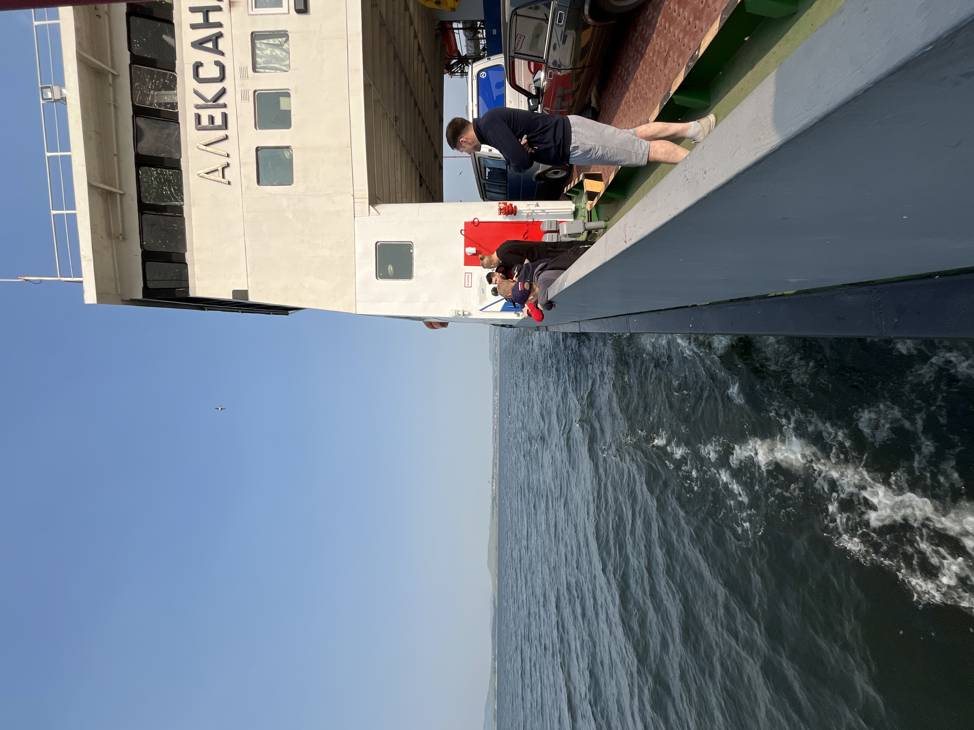 The image captures a view from the deck of a ferry traveling on a body of water, likely the Krasnoyarsk Reservoir, near the village of Novoselovo, Russia. The ferry has the Cyrillic name "АЛЕКСАНДР" visible on its side. Several people are standing on the ferry's deck, some facing the water and others conversing. A dark-colored car is parked on the ferry's deck, along with what appears to be a white van or bus. The water is choppy, indicating the ferry is in motion, and a wake is visible trailing behind. The sky is clear and blue, with a distant shoreline featuring hills and some buildings, consistent with the landscape around Novoselovo. The overall scene suggests a mode of transportation and leisure activity common in the region.
