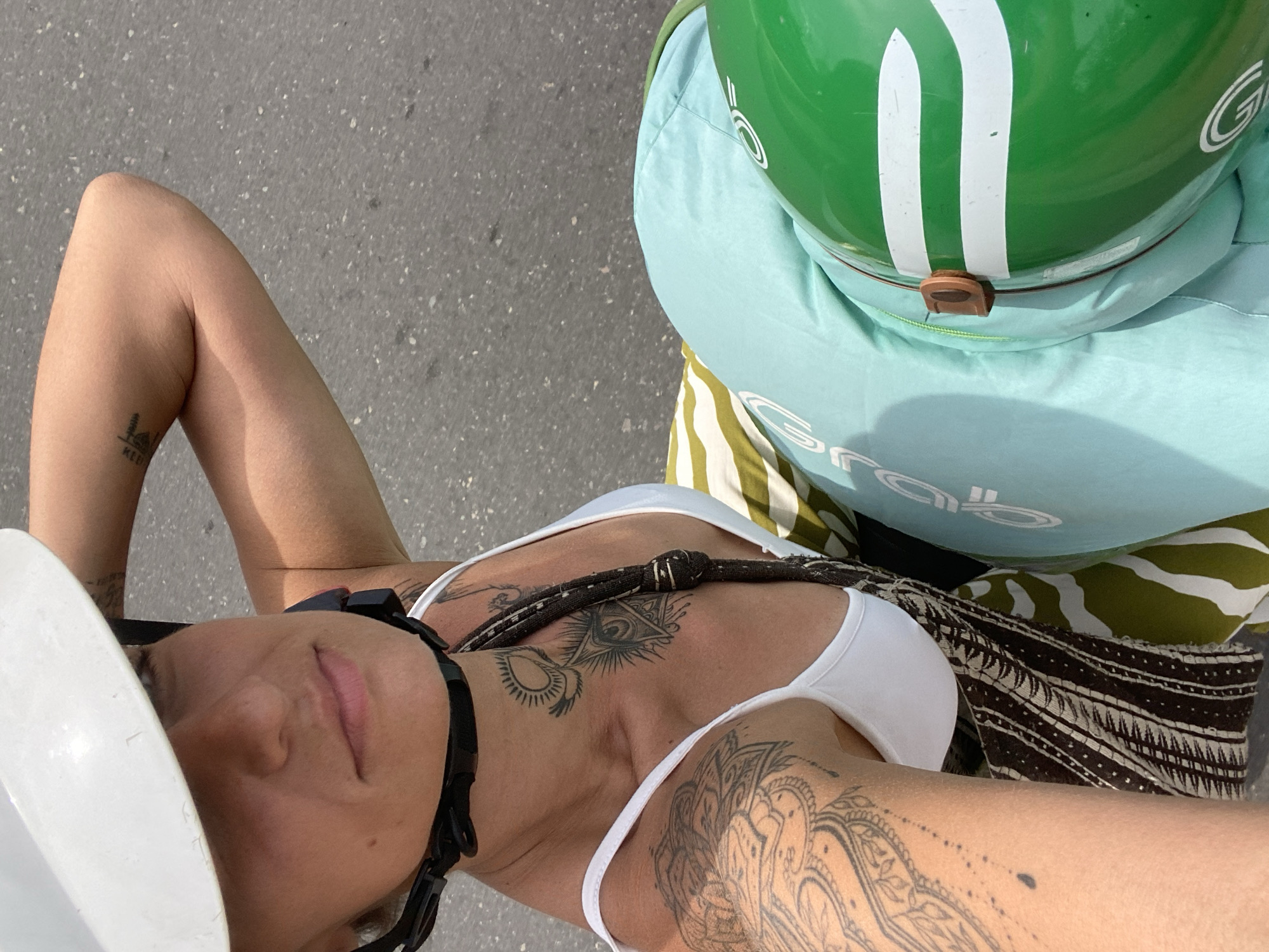 A first-person perspective shot shows a person wearing a white helmet and a white tank top with intricate tattoos on their arm and chest. They are riding what appears to be a motorbike on an asphalt road in Phu Thuy Ward, Vietnam. The person's arm is bent, and their hand is on the helmet. A light blue fabric is visible, partially covering a green helmet with white stripes. The background is a gray asphalt road with small white speckles.