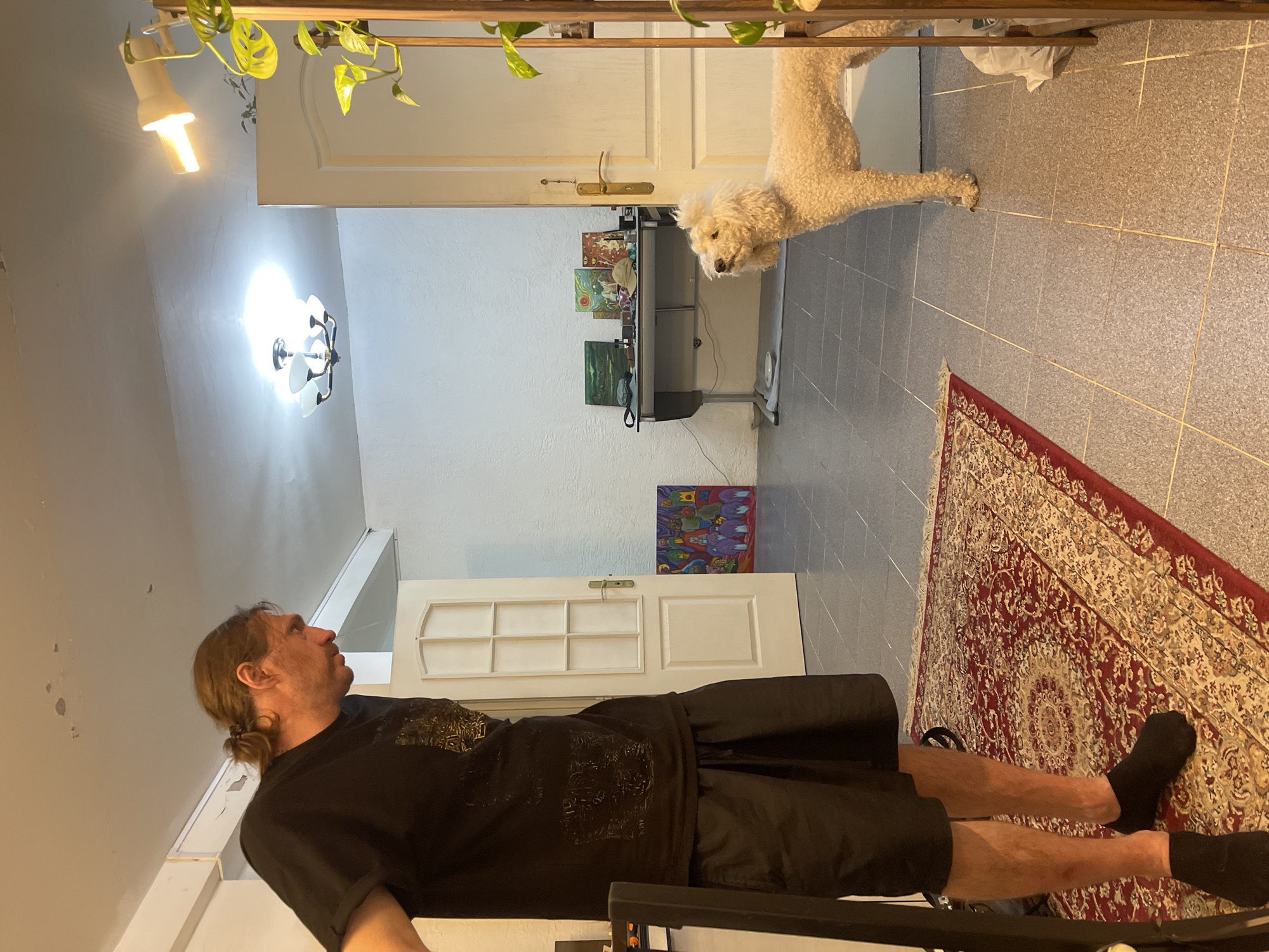 A man with a ponytail, wearing a black t-shirt and black shorts, stands in a room, looking down and to his right. To his right, a white poodle stands near a partially open door, looking up at the man. The room has tiled floors and white walls. A red oriental rug is on the floor in the foreground. In the background, a desk with several framed pictures and a baseball cap sits against the wall. A ceiling light fixture is visible. A floor lamp with a bright bulb is also present. The scene appears to be indoors, in what might be a home or office. There is no indication that this image is related to Krasnoyarsk, Russia.