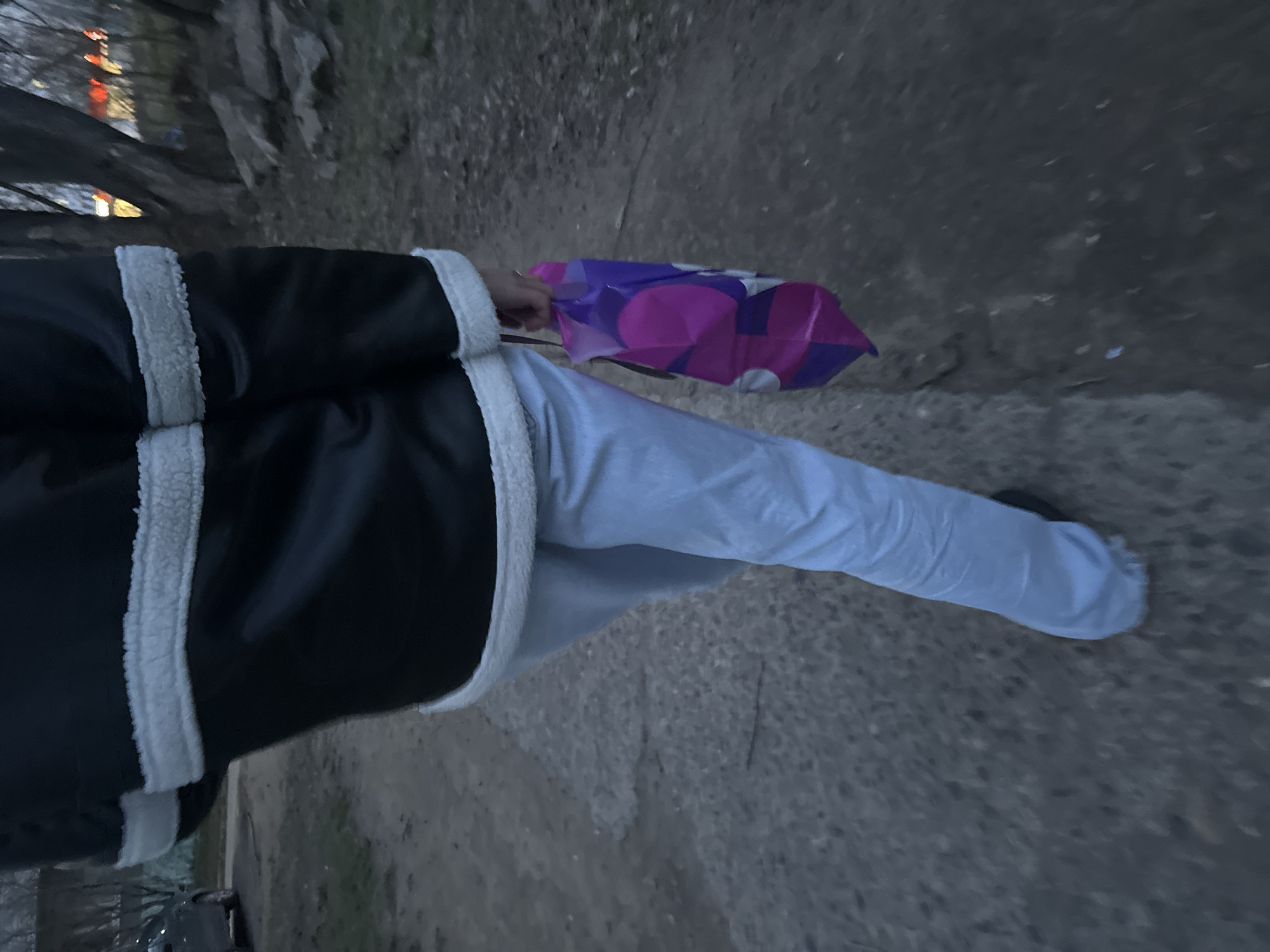 The image, taken in low light and oriented sideways, depicts a person from the waist down, walking or standing on an outdoor path. The individual is wearing a dark, possibly black, jacket with a contrasting white, fluffy or shearling-like lining visible along the edges and sleeves. They are also wearing light-colored, likely white or light grey, pants that appear somewhat wrinkled.
The person is holding a vibrant purple and pink plastic bag in their right hand. The bag features a repeating geometric pattern composed of overlapping circular or oval shapes, with some sections colored purple and others pink. There is no clearly legible text or brand logo visible on the bag.
The scene is an outdoor setting, with the ground appearing to be a dark, rough path, possibly asphalt or dirt. In the background, out of focus, are dark trees or bushes. Several distant lights, appearing orange and white, are faintly visible through the foliage, suggesting the time of day is either dusk or nighttime. The overall atmosphere is dim, indicating late evening or early morning. The setting appears to be a natural or semi-urban outdoor area, possibly near a park or residential street.