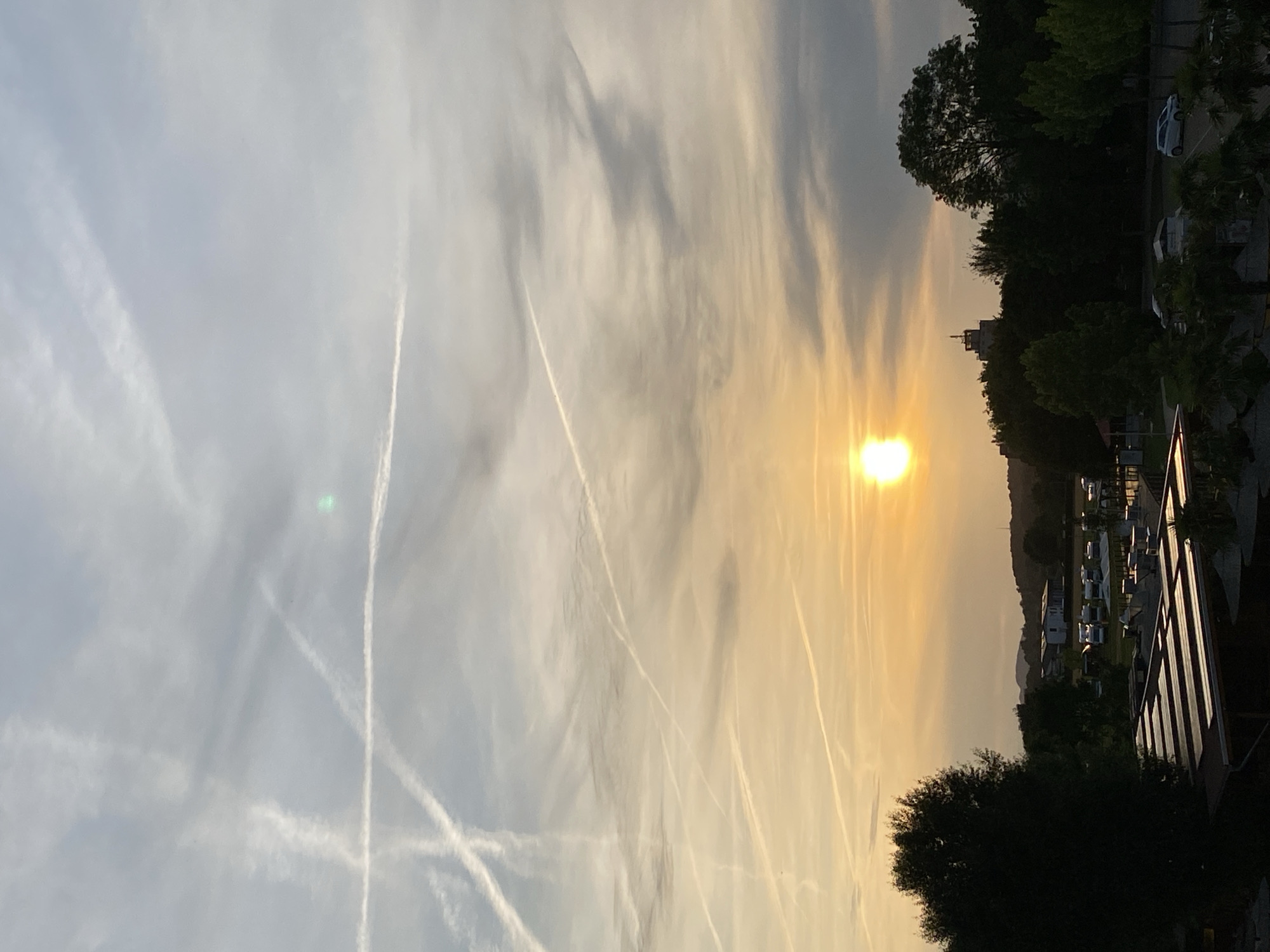 The image depicts a sunset scene. The sun is visible in the upper central portion of the frame, casting a warm orange glow across the sky, which is filled with numerous contrails from aircraft, creating a complex pattern of white lines against the muted blue and grey clouds. In the foreground, silhouetted trees and structures are visible. The landscape below shows a collection of recreational vehicles or caravans parked in a field, with some buildings and a distant tower visible on the horizon. The overall atmosphere is serene, with the setting sun dominating the sky. The location is identified as Rinteln, Germany.