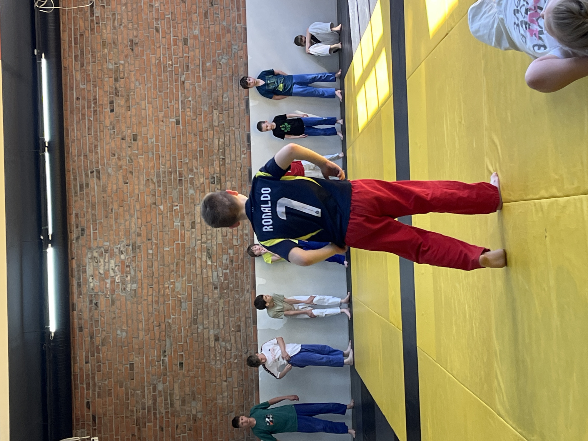 A group of children are practicing judo in a gymnasium. The gymnasium has a brick wall, and the floor is covered with yellow mats. The children are lined up in rows, and some are wearing judo uniforms, while others are wearing street clothes. One child is wearing a "Ronaldo 7" jersey. The location is likely in Krasnoyarsk, Russia.