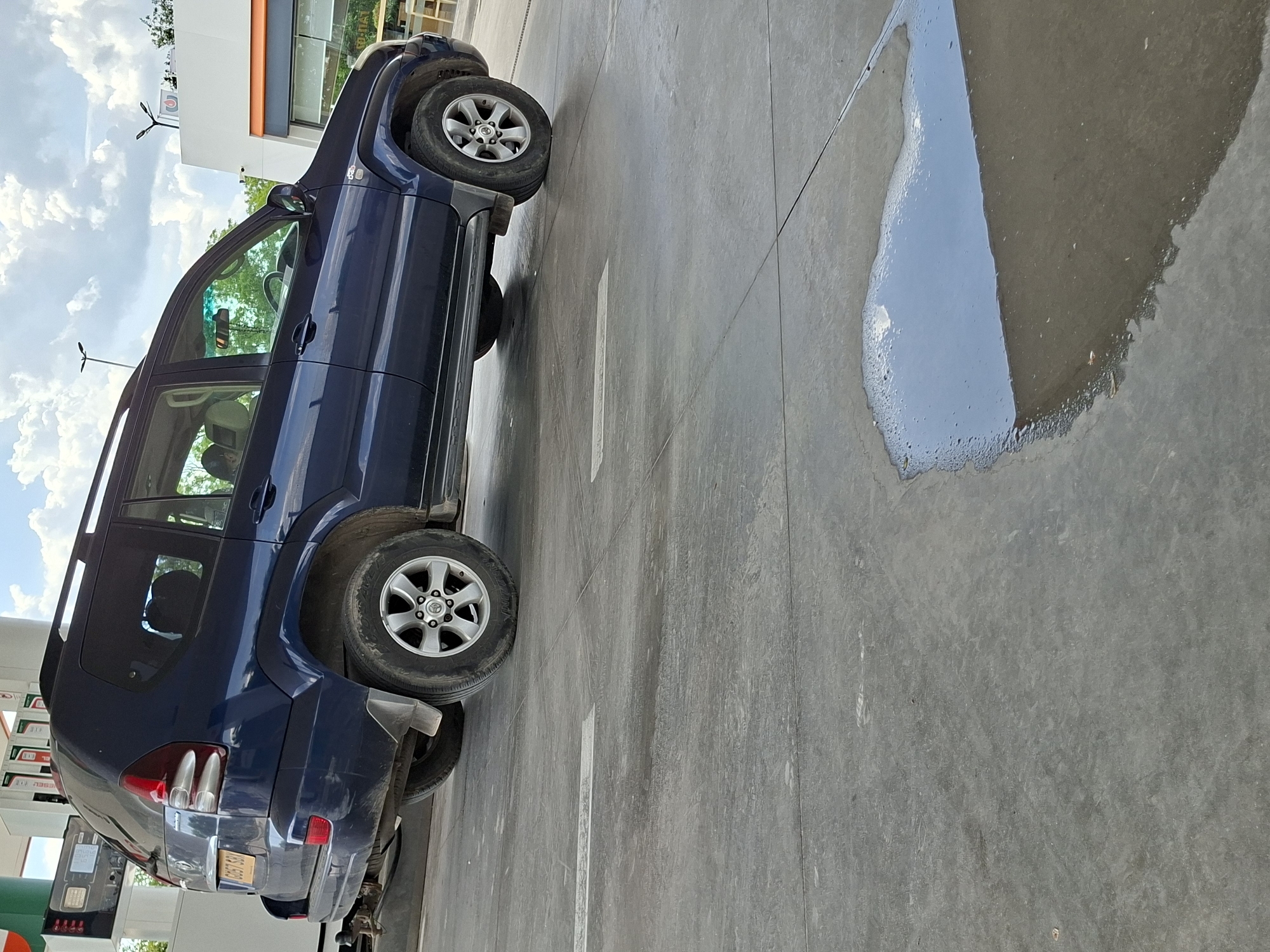 The image shows a dark blue Toyota Land Cruiser parked at a gas station in Plovdiv, Bulgaria. A puddle of spilled fluid reflects the sky. The station's canopy is visible above the vehicle. The ground is gray concrete with white lane markings.