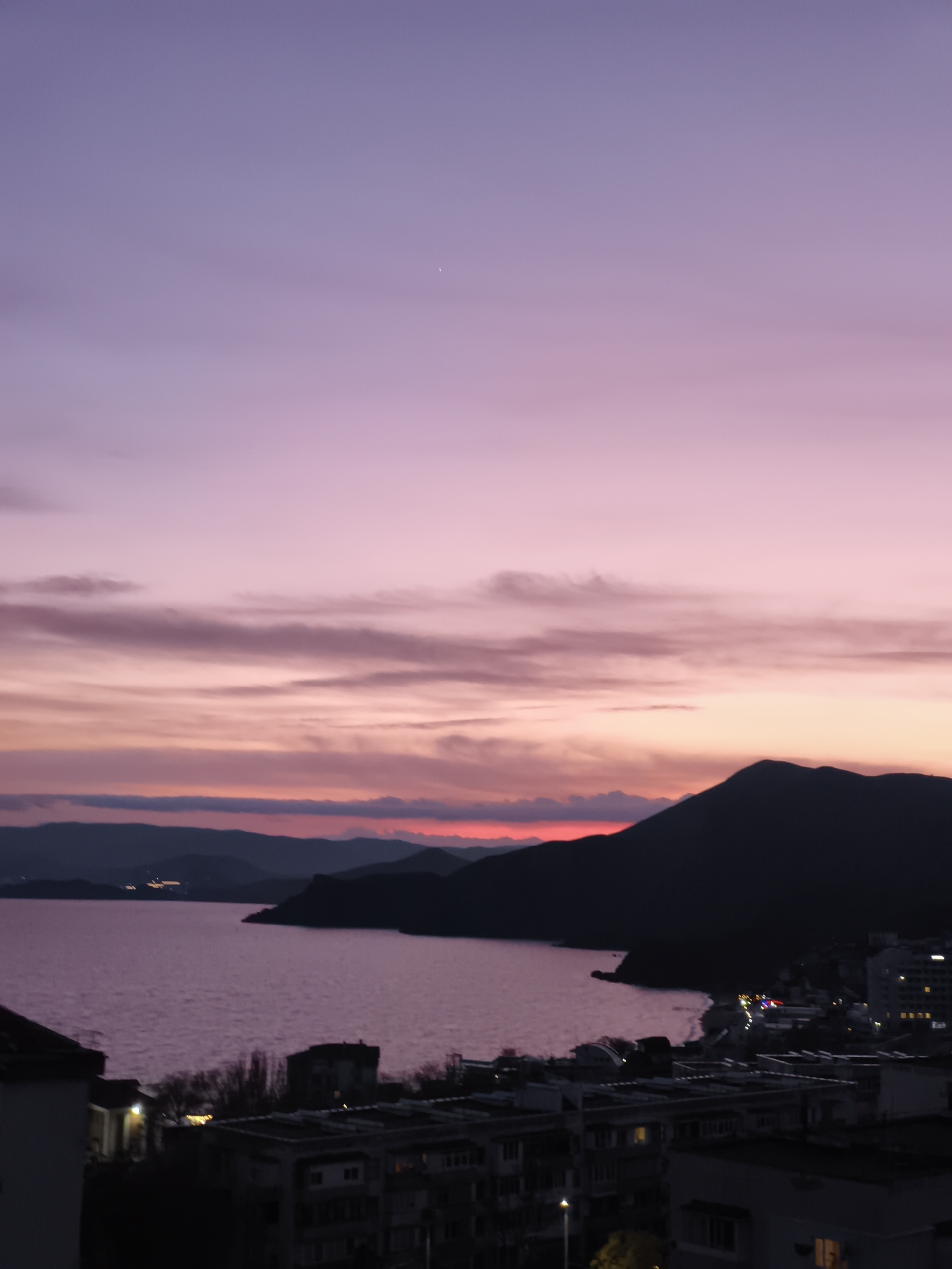 A scenic view captures a city situated alongside a body of water, likely the sea, under a twilight sky. In the foreground, the city's architecture is visible, comprising apartment blocks and buildings with illuminated windows, indicating the presence of residents. The waterfront is softly illuminated, with scattered lights reflecting on the water's surface.
In the middle ground, the sea extends, bordered by a landmass or peninsula that juts out, silhouetted against the fading light of the sunset. The sky transitions from a deep purple at the zenith to a warmer, reddish hue near the horizon, punctuated by scattered clouds that catch the last rays of the sun.
The image suggests a tranquil evening in the city, with the natural beauty of the sunset contrasting against the urban environment. The location is speculated to be within the vicinity of the Орджоникидзевский поселковый совет region of Ukraine, based on known landmarks and characteristics.