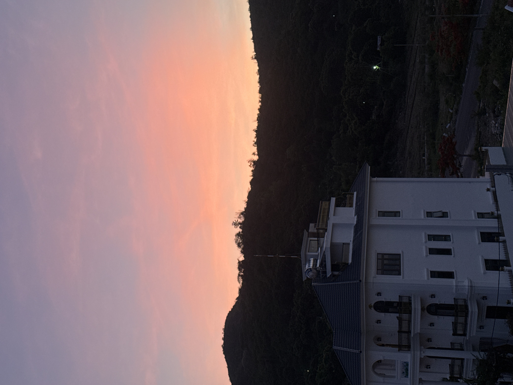 A wide shot captures a villa at dusk, silhouetted against a gradient sky of purple and orange, with a dark, forested hill in the background. The villa, painted white, features multiple stories, balconies with dark railings, and arched windows. A water tank and antenna are visible on the roof. The surrounding area appears to be a residential development, with a paved road leading past the property and some scattered red-flowering trees. The lighting suggests it is either dawn or dusk.