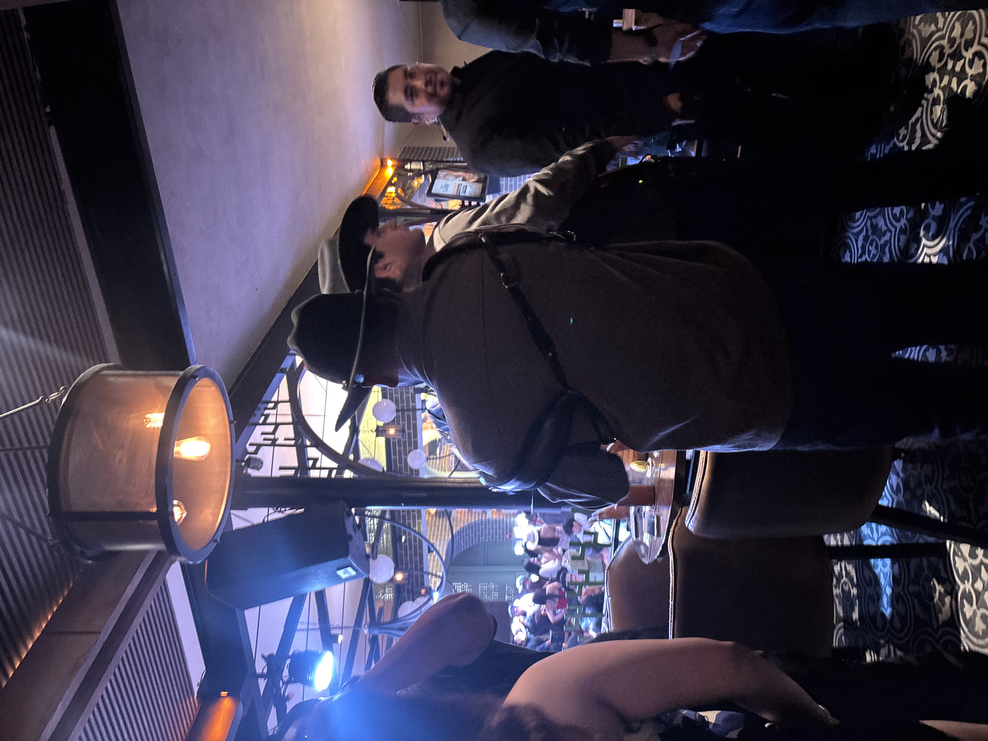 The image depicts an indoor scene in a possibly upscale establishment, likely a bar or restaurant. There are people present, some standing and wearing hats, and others seated at what seem to be bar stools. The lighting is a mix of incandescent from a cylindrical hanging lamp and blue from an unseen source. The bar appears to have a view of a public area with more people, visible through a structural frame.
Given the context of Puebla, Mexico, and the architecture visible through the structure, one can speculate that the location is overlooking or situated within a central historic area in the city.