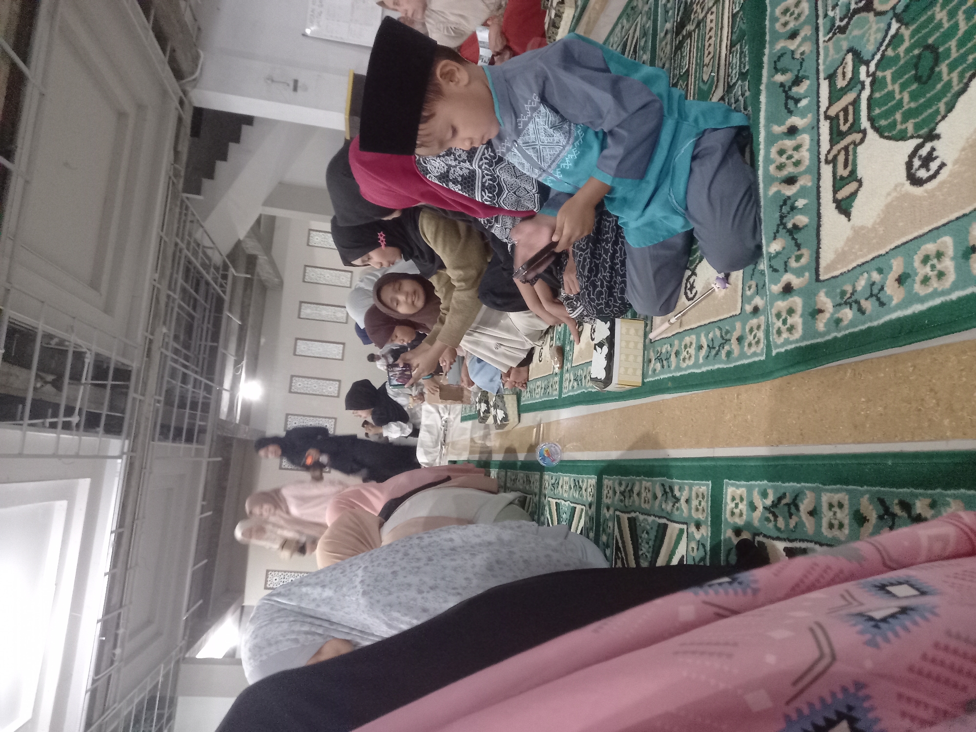 In Banyumulek, Indonesia, a group of people, predominantly women and children, are gathered indoors, seated on green carpeted prayer mats. The room has a ceiling with exposed industrial-style framing and lighting. The individuals are facing generally towards the right side of the frame, with most dressed in traditional Islamic attire, including hijabs and modest clothing. Some are looking at their mobile phones, while others appear to be engaged in conversation or observing something off-camera. A young boy in the foreground is wearing a peci hat and a teal and grey outfit, holding a mobile phone. The prayer mats have intricate patterns in various shades of green, white, and beige. Boxes are placed on the mats, some appearing to be tissue boxes. The overall atmosphere suggests a communal gathering or religious event.