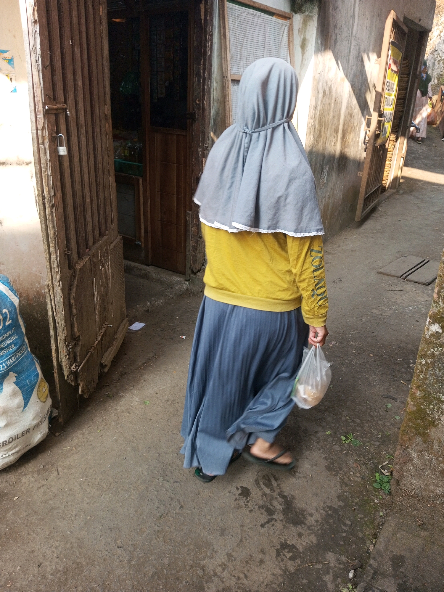A person wearing a gray hijab and a yellow long-sleeved shirt is walking away from the camera, holding a transparent plastic bag containing items. They are also wearing a blue, pleated skirt and black sandals. The person is walking on a dirt path beside a building. To the left of the person is a large, blue and white bag with text in Indonesian. Further to the left, a wooden gate with a padlock is visible. The environment appears to be an outdoor urban setting.