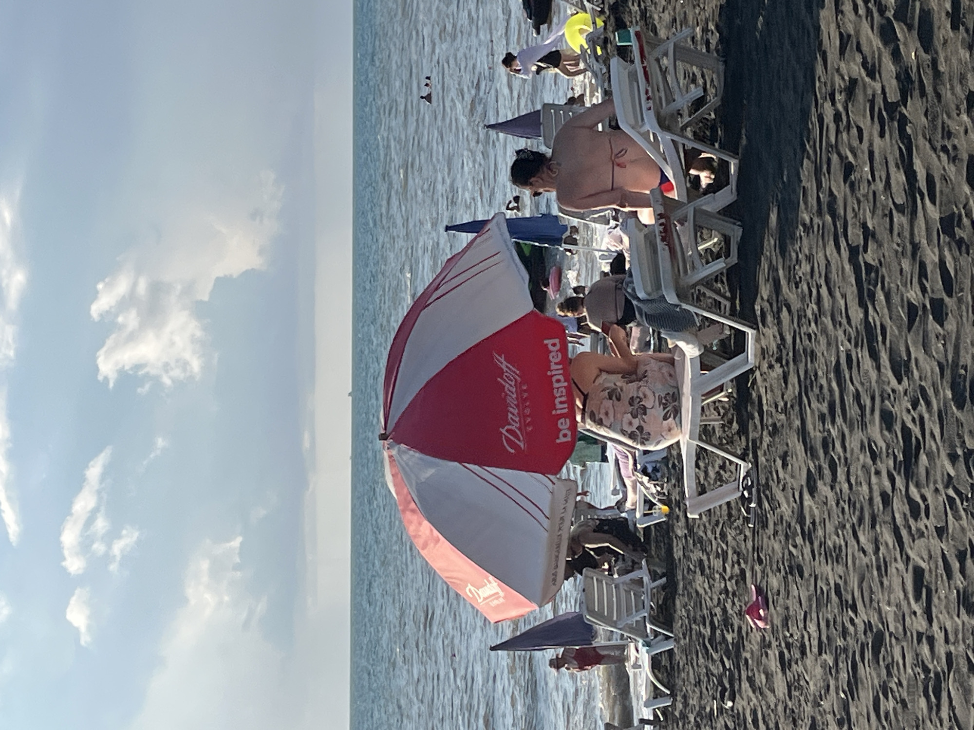 This photograph captures a scene on the beach in Ureki, Georgia, a location known for its magnetic sand. In the foreground, dark sand stretches across the image, with several white beach chairs scattered on it. A pink flip-flop lies on the sand to the left. Dominating the mid-ground is a large, red and white striped umbrella with the "Davidoff" logo visible. Underneath this umbrella, several people are seated on chairs, some in swimwear, with their backs to the camera. A woman in a floral dress is in the immediate foreground, sitting in a beach chair. To the left, another umbrella, a dark blue one, is visible, and further back, a woman in a red swimsuit stands in the shallow water. In the background, the sea stretches out to the horizon, with a few small boats or distant figures visible on the water. The sky above is partly cloudy, with patches of blue sky and white, fluffy clouds. The overall impression is a relaxed beach scene on a sunny day.