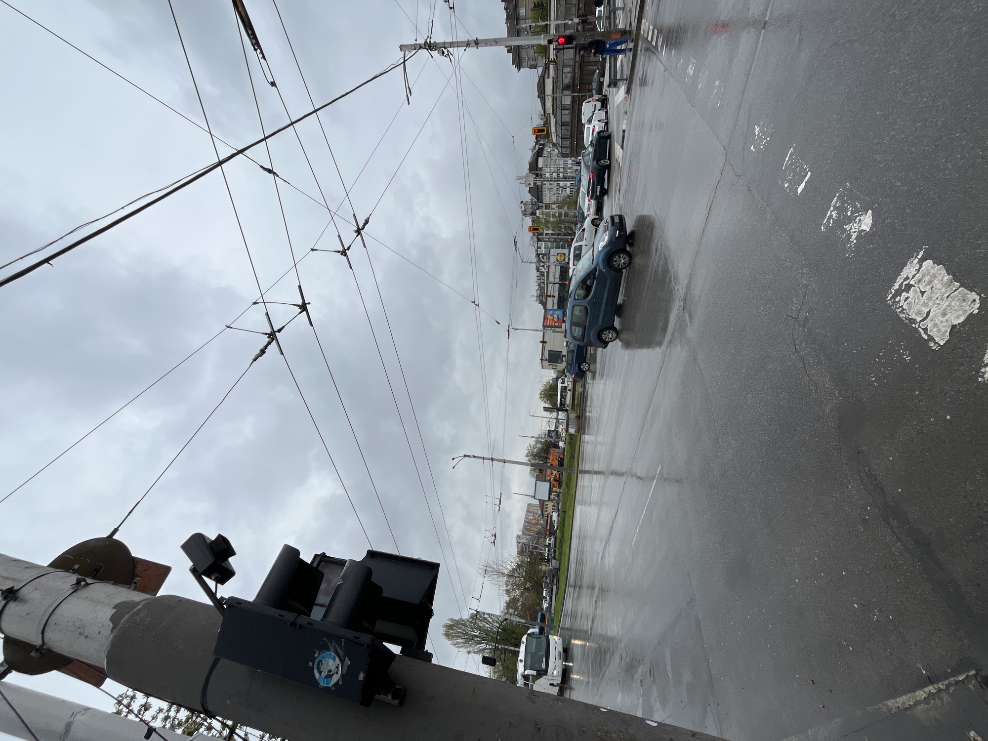 The image displays an urban street scene on an overcast day, characterized by a wet asphalt road, indicating recent rain. Overhead wires, characteristic of trolleybus or tram systems, crisscross the cloudy sky.
In the foreground, a large concrete utility pole with traffic signals is prominently featured on the left. One visible traffic light displays a red signal. A circular blue and white logo sticker is affixed to the pole, showing a stylized white letter (resembling 'C', 'G', or 'S') within a blue circle, encompassed by a white ring that evokes a lifebuoy. Below this central emblem, some small, unreadable text is present.
Further down the street, multiple vehicles are visible, appearing either stopped or moving slowly in traffic. A blue minivan is prominent among the cars. Another red traffic light is seen in the mid-ground, reinforcing the sense of halted movement. On the far right, a person stands on the sidewalk. The background features several multi-story buildings lining the street, and sparse trees are visible. Faded white markings are painted on the wet road surface. A white bus-like vehicle is also discernible in the distance. The overall scene depicts a typical city street environment under damp, cloudy conditions.