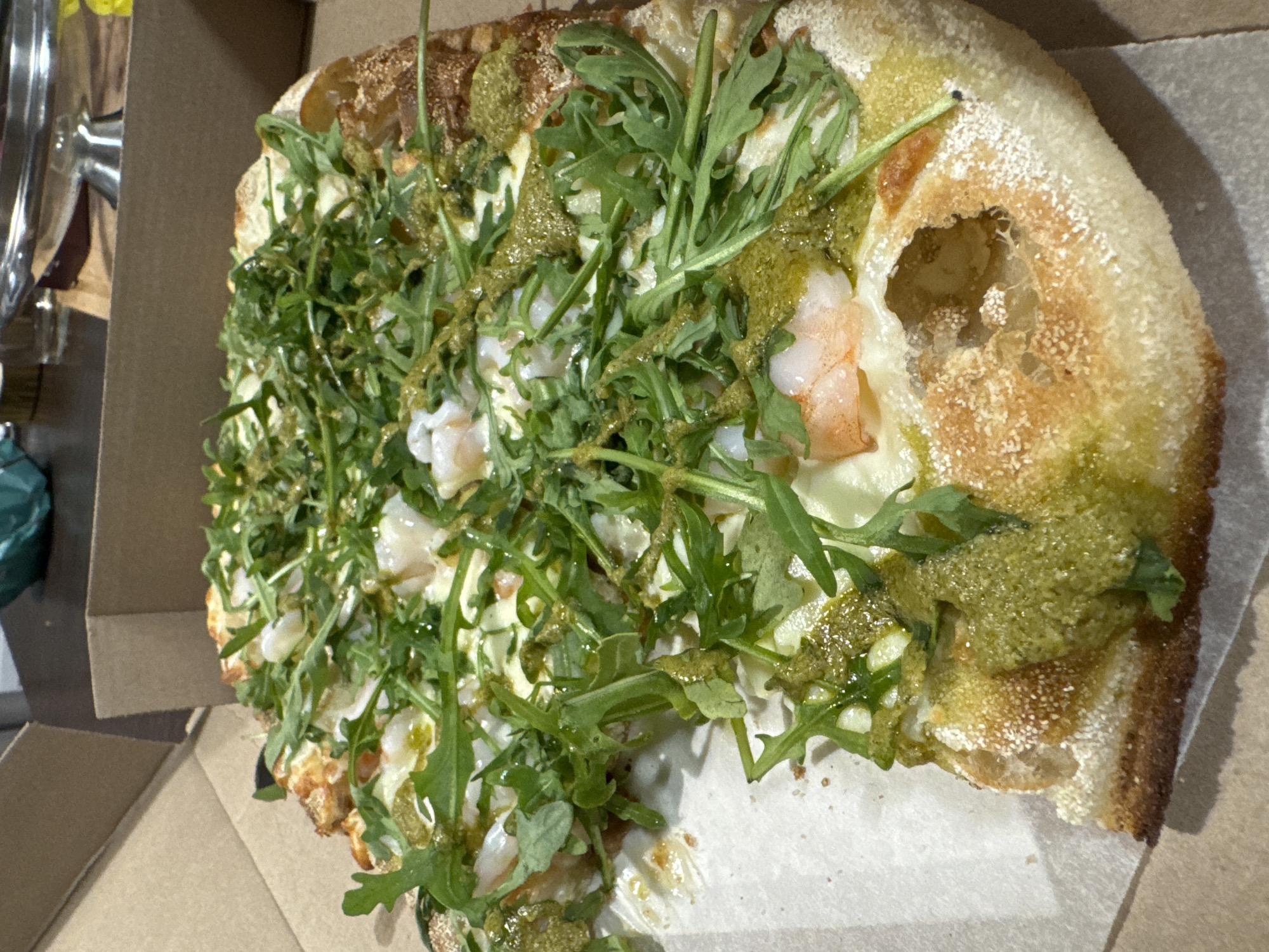 The image shows a partial view of a rectangular pizza inside a cardboard box. The pizza has a thick crust, with a hole in one section revealing the interior. The toppings include what appears to be arugula, some sort of green pesto-like sauce, and shrimp. There are no people visible. The environment appears to be indoors. The image provides no information that would suggest Izhevsk, Russia as a location.