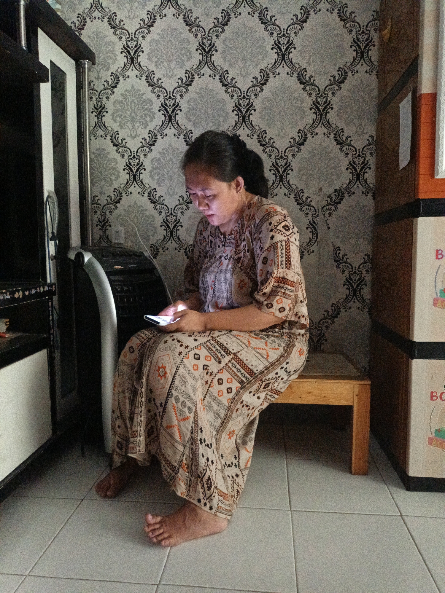 In Cilegon, Indonesia, a woman sits on a wooden bench, engrossed in her smartphone. The room is decorated with a grey and black damask wallpaper. To her left, a tall, dark cabinet with a white section is visible, alongside a silver and black air cooler. To her right, stacked boxes, one with "BO" printed on it, form part of a storage unit. The floor is tiled in a light grey. The woman wears a long, patterned dress in shades of beige, brown, and orange. Her feet are bare and visible beneath the hem of her dress.