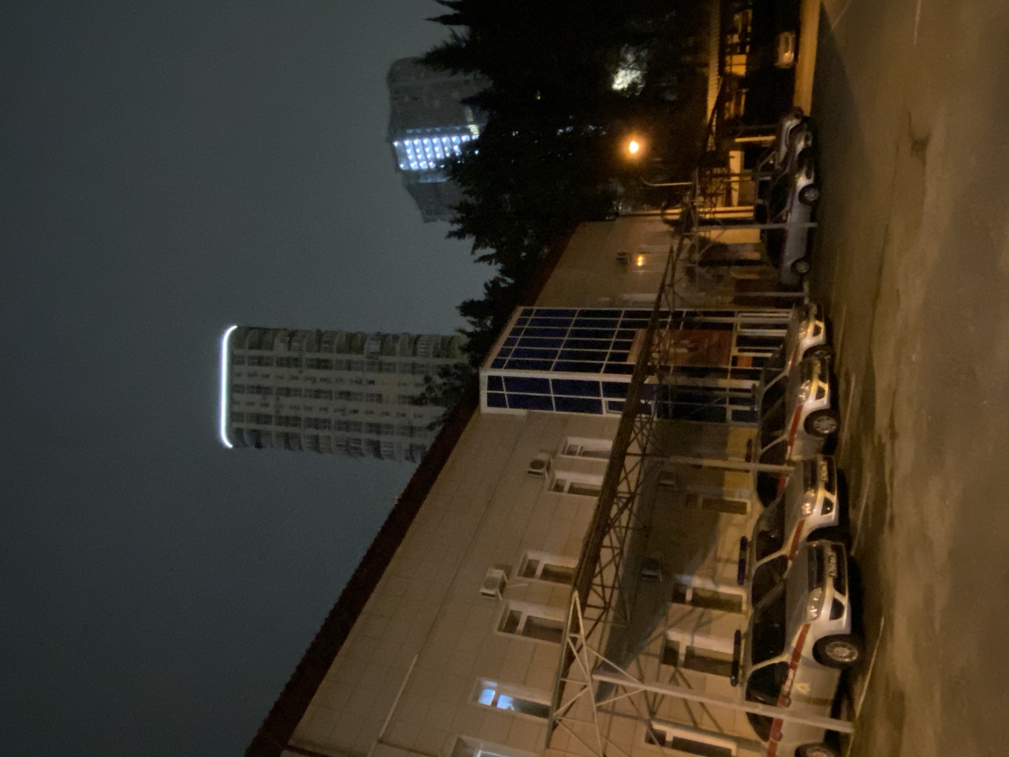 The image depicts a nighttime scene in what appears to be a parking lot or area adjacent to a building, possibly in Sochi, Russia. Multiple vehicles, some appearing to be emergency or official cars with light bars on their roofs, are parked in a row. Overhead structures are visible, possibly part of a covered parking area or building support. In the background, there are high-rise buildings lit up at night. The sky is dark and overcast.
