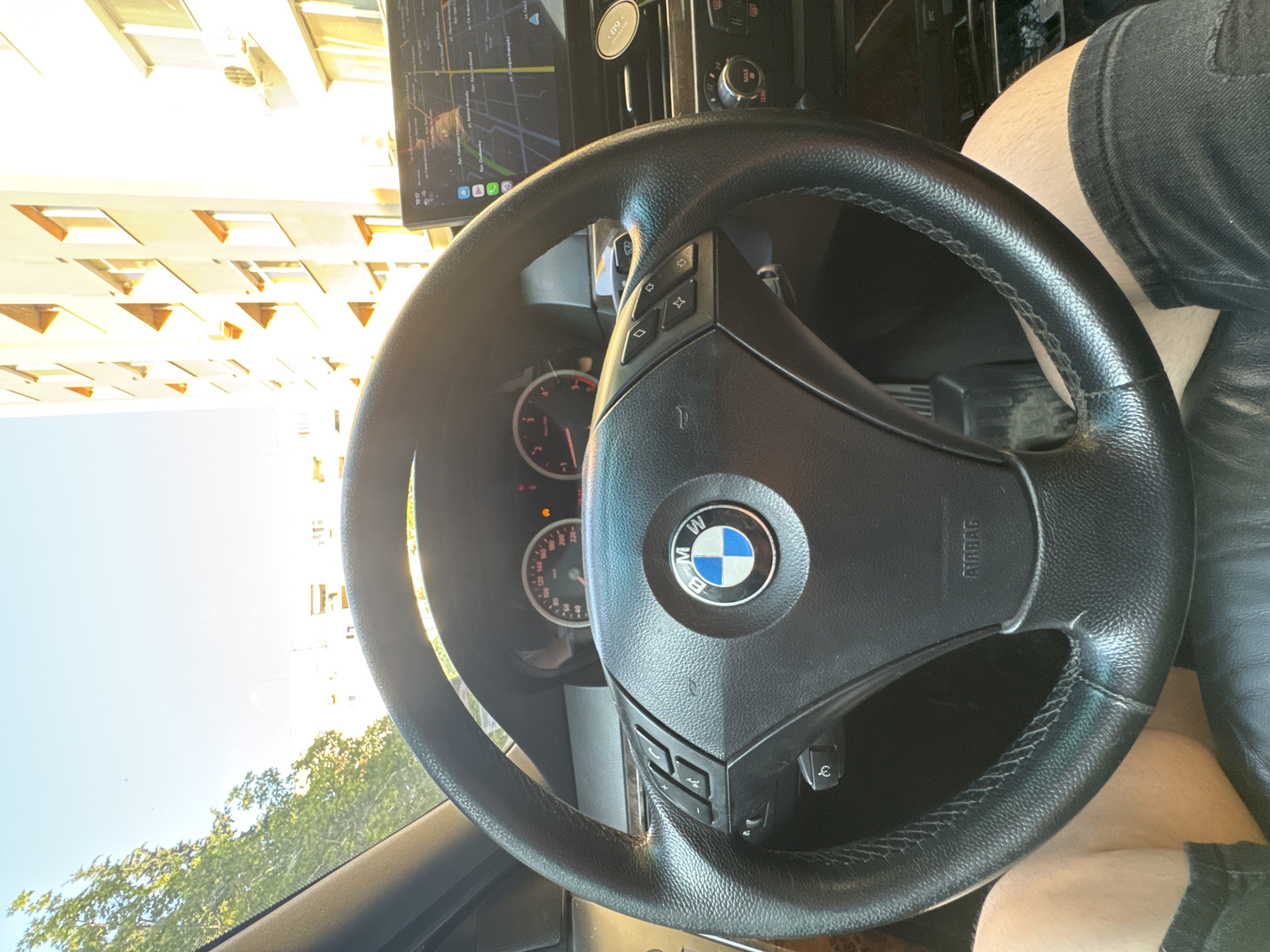 The image depicts the interior of a stationary BMW vehicle, likely an E60/E61 5-series, on a sunny day in Stara Zagora, Bulgaria.
In the immediate foreground, the lower thighs and bare knees of a person, likely the driver, are visible, suggesting the person is wearing shorts. The prominent object is a black leather-wrapped three-spoke BMW steering wheel with visible stitching and signs of wear. The central airbag cover features the blue and white BMW emblem. The left side of the steering wheel contains control buttons for phone functions (answer, end call, voice command) and volume. The right side contains diamond-shaped buttons, likely for cruise control or media navigation.
Behind the steering wheel, the vehicle's instrument cluster is visible, showing a speedometer with the needle at 0 km/h and a tachometer with the needle at 0 RPM, indicating the car is not in motion. Several warning lights are illuminated on the dashboard. Above the central dashboard, an aftermarket infotainment screen is mounted, displaying a map application (resembling Google Maps) with street names written in Cyrillic. Visible street names include "ул. Генерал Гурко" (General Gurko Street), "ул. Стефан Караджа" (Stefan Karadzha Street), "ул. Славянска" (Slavyanska Street), "бул. Руски" (Ruski Boulevard), and "ул. Цар Симеон Велики" (Tsar Simeon Veliki Street), all confirming the location as Stara Zagora, Bulgaria. The time displayed on the screen is 10:17. Below the infotainment screen, parts of the vehicle's climate control system are visible, showing dials with "MAX" and "TEMP 22" settings. A small black circular object with "09." is affixed to the dashboard to the right of the climate controls.
Through the windshield, the bright blue sky and lush green trees are visible on the left. On the right, a multi-story building with a light-colored facade and multiple windows with brown or orange frames is illuminated by direct sunlight, causing significant glare. An external air conditioning unit is mounted on the side of this building.